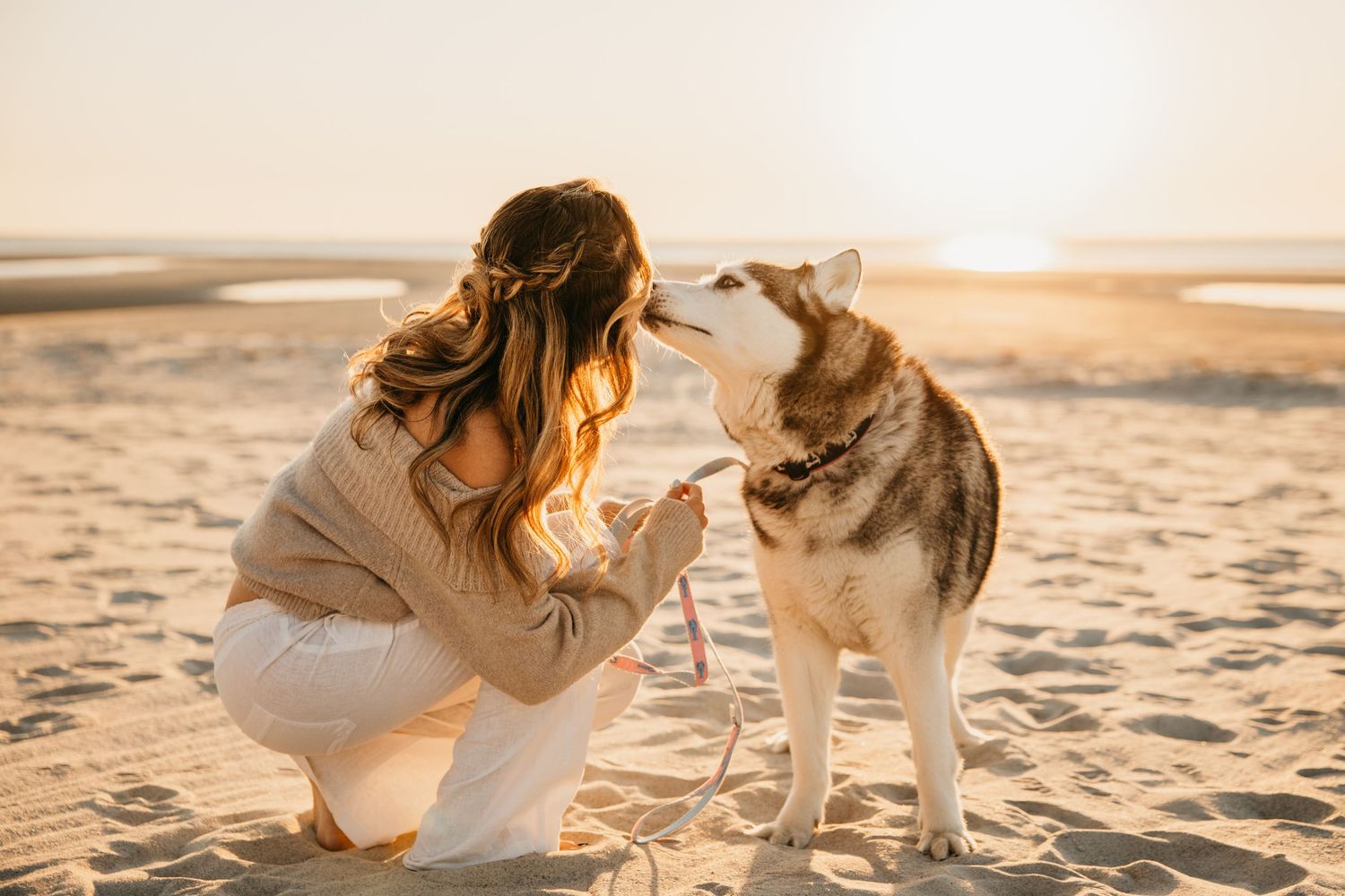 Woman in beige sweater kneeling on sandy beach, sharing a tender moment with a Husky dog at golden sunset.
