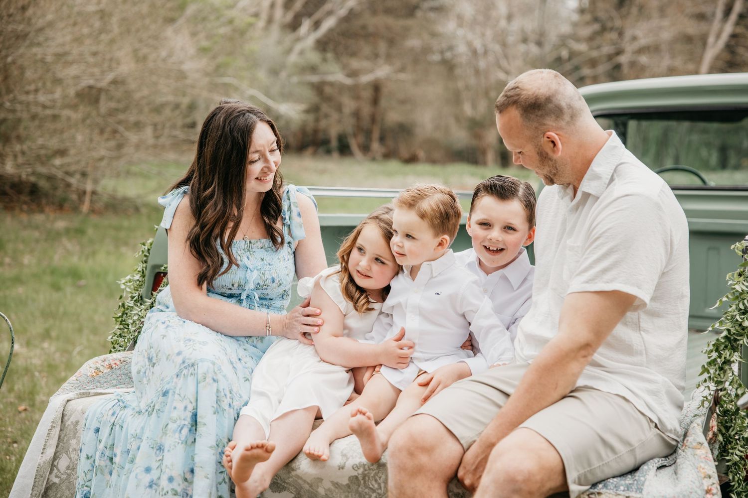 Family of five sitting on vintage truck tailgate outdoors, parents smiling at three young children in spring garden setting.