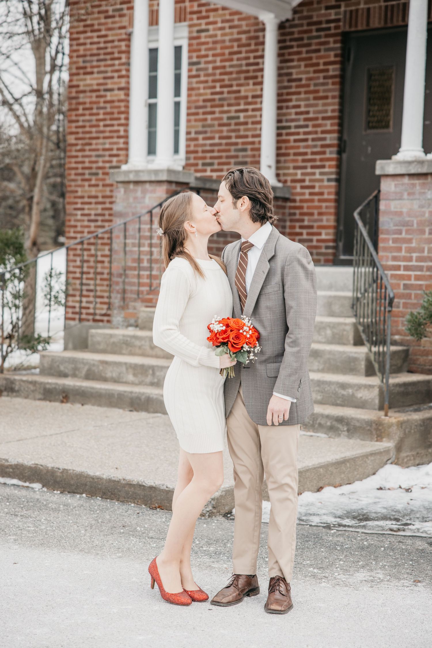 Couple shares romantic moment in front of brick building with red floral accents.