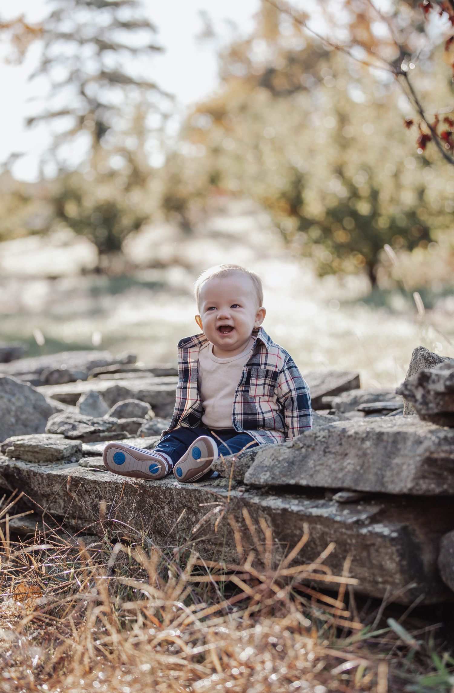 A smiling baby in plaid shirt sits on stone wall during autumn.
