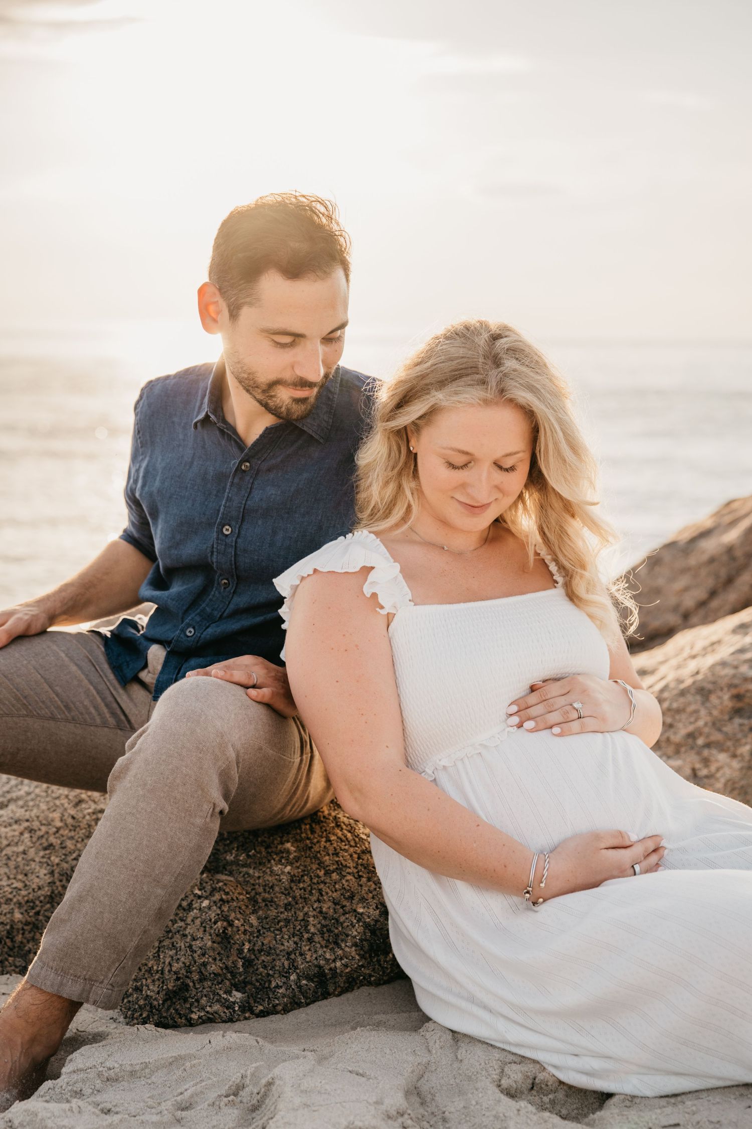 Couple sitting on rocks by the ocean at sunset, pregnant woman in white dress cradling baby bump with partner beside her.