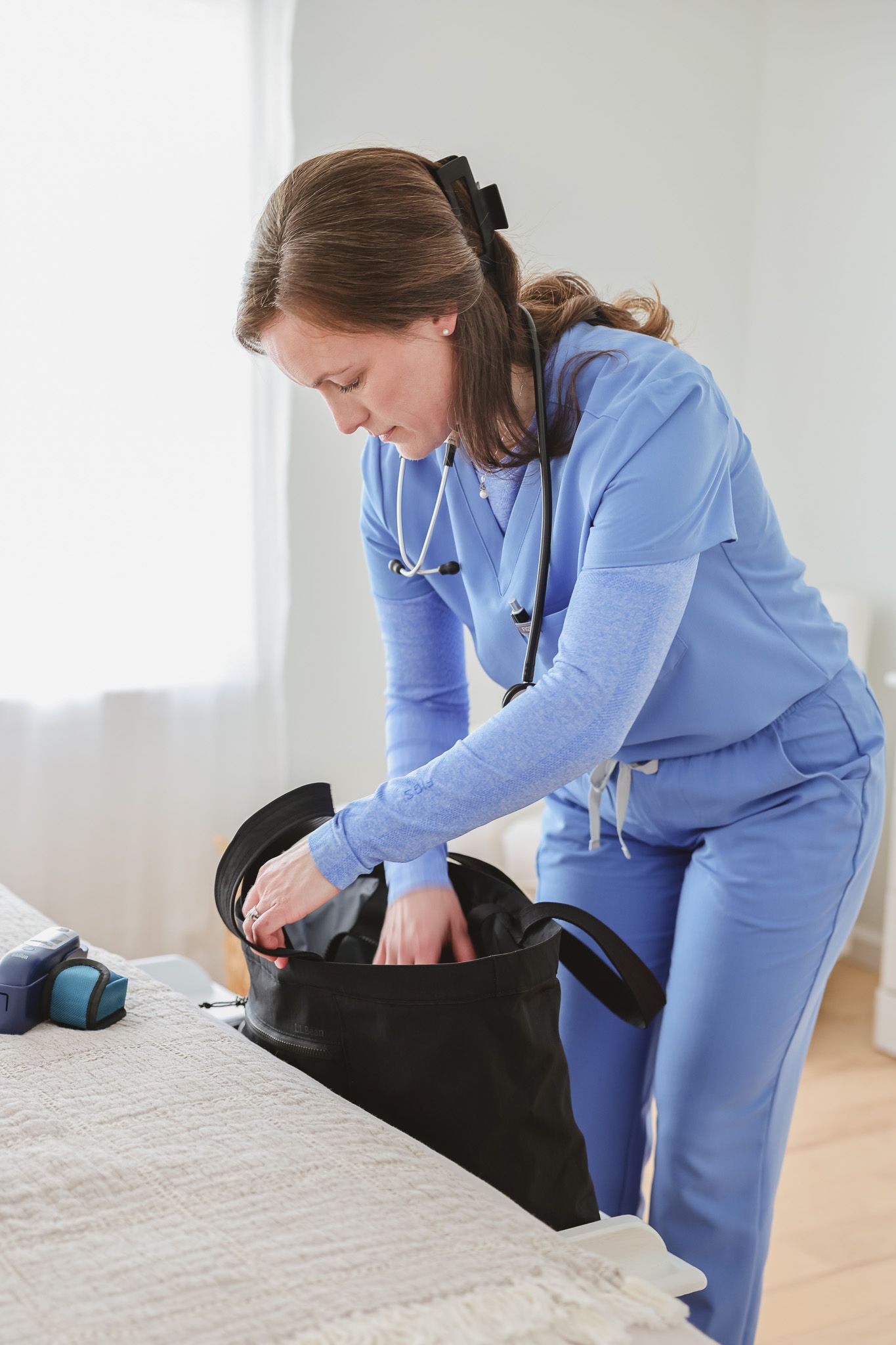 Female nurse in blue scrubs with stethoscope unpacking medical bag on table during home health visit.