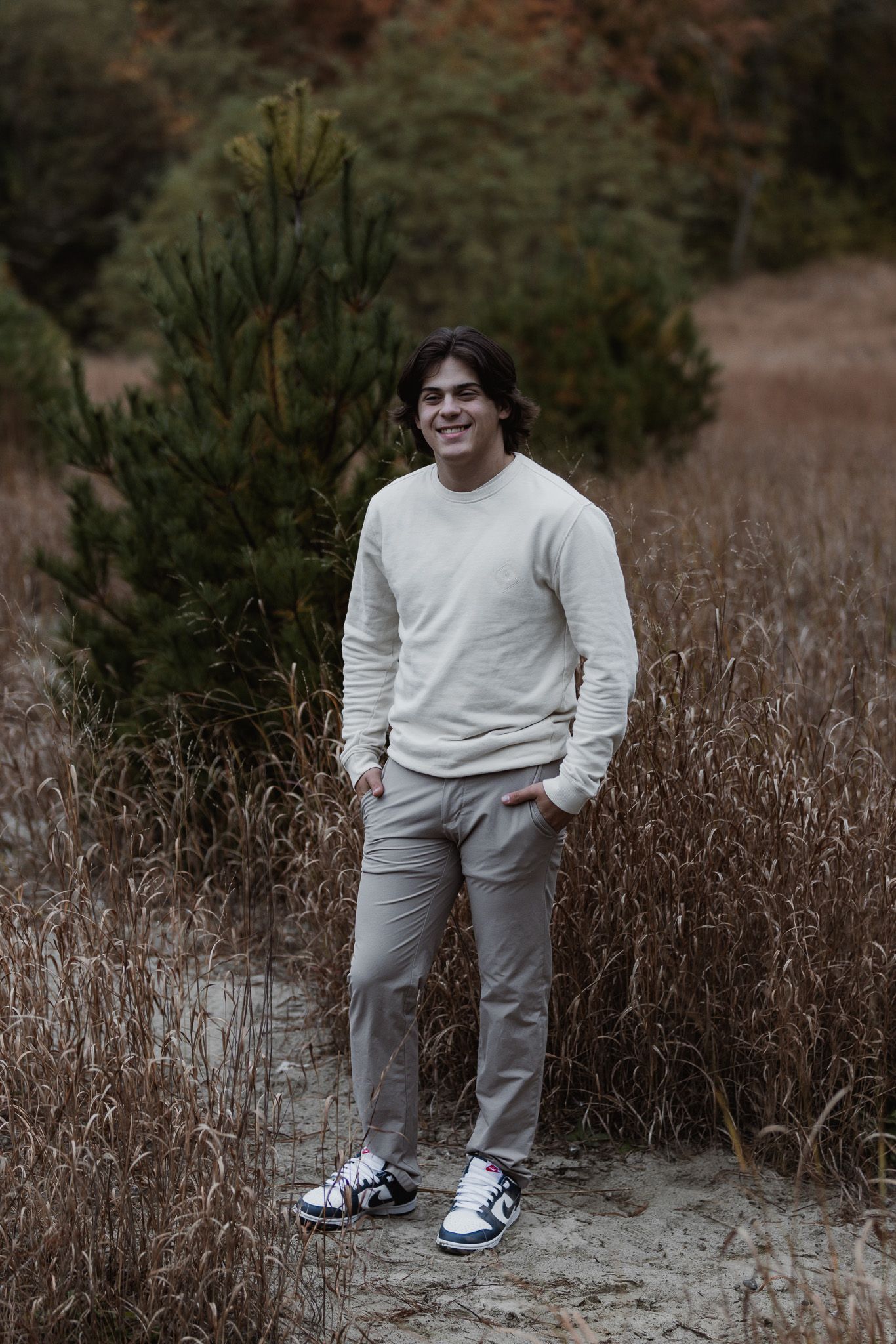 Person in white sweater and gray pants stands among dried grass and pine trees.