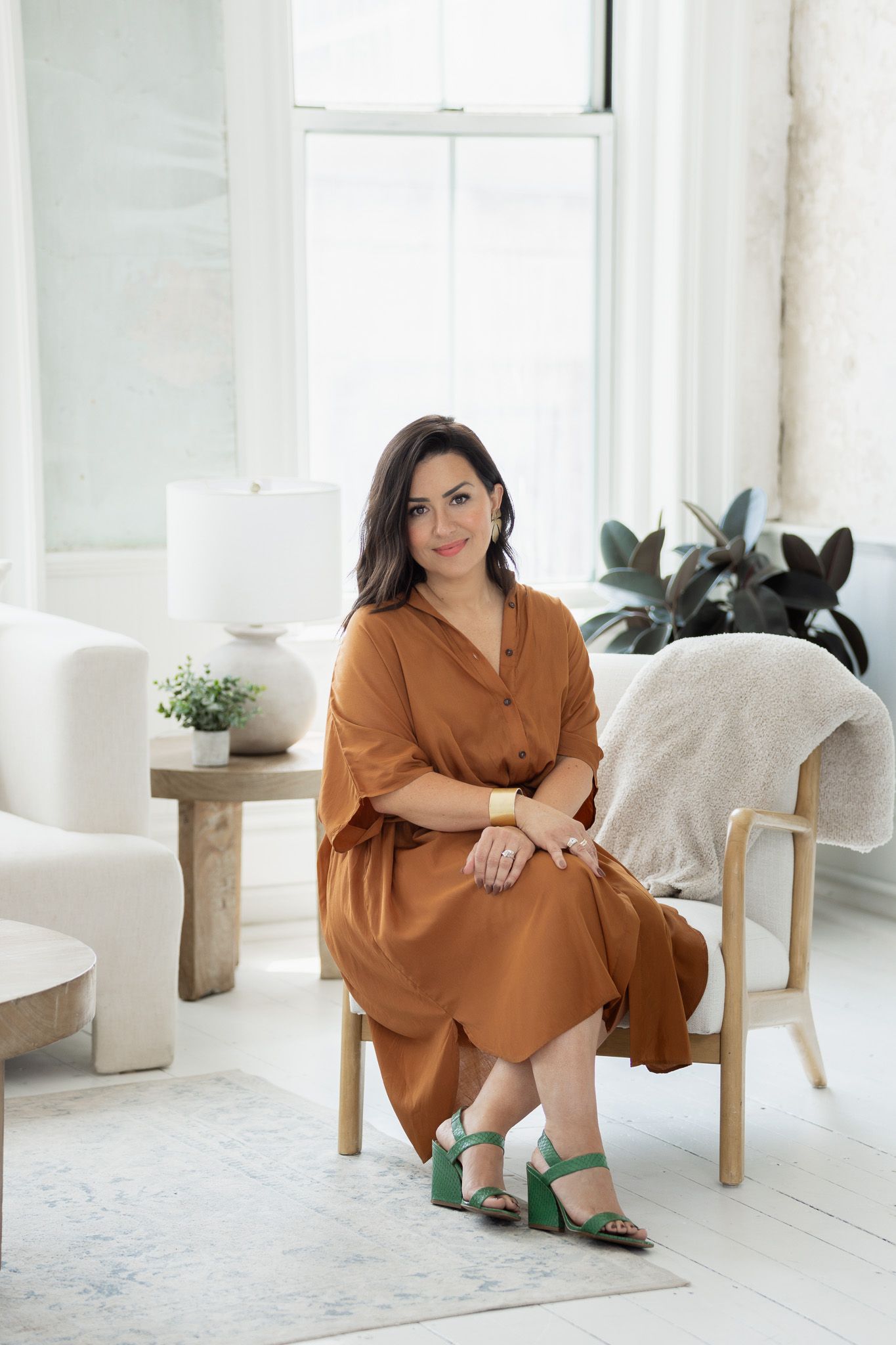 Person in rust-colored dress sits elegantly on a wooden chair in a minimalist white room.