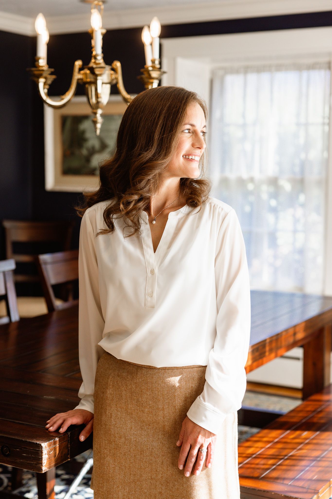 Professional in white blouse and tan skirt poses in an elegant dining room with chandelier.
