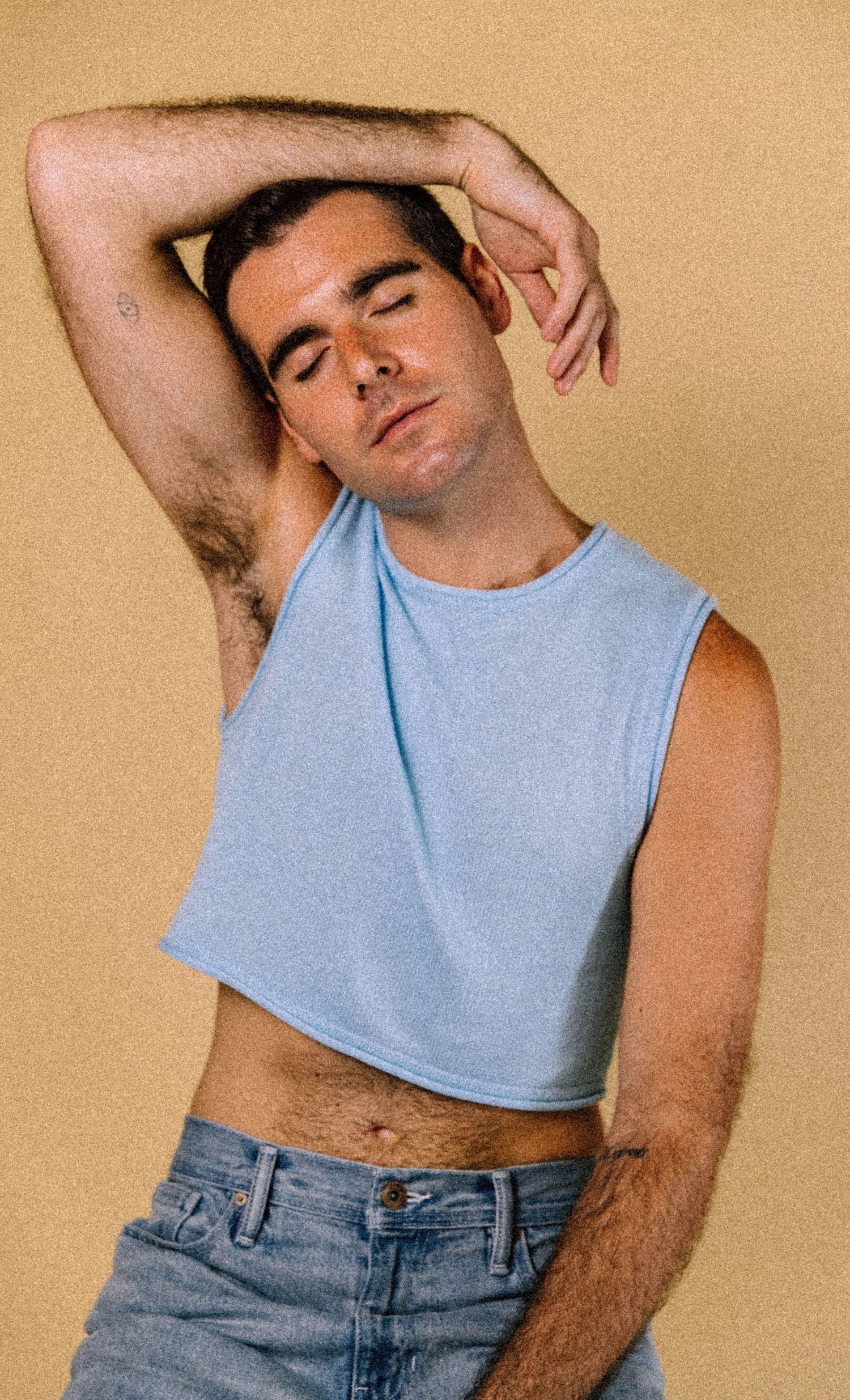 Fashion poses in a cropped light blue tank top and denim against a beige background.