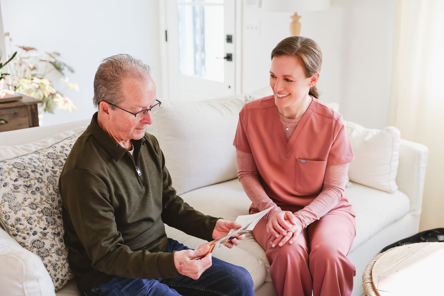 A smiling nurse in pink scrubs reviews documents with an elderly man on a white sofa during a home care visit.
