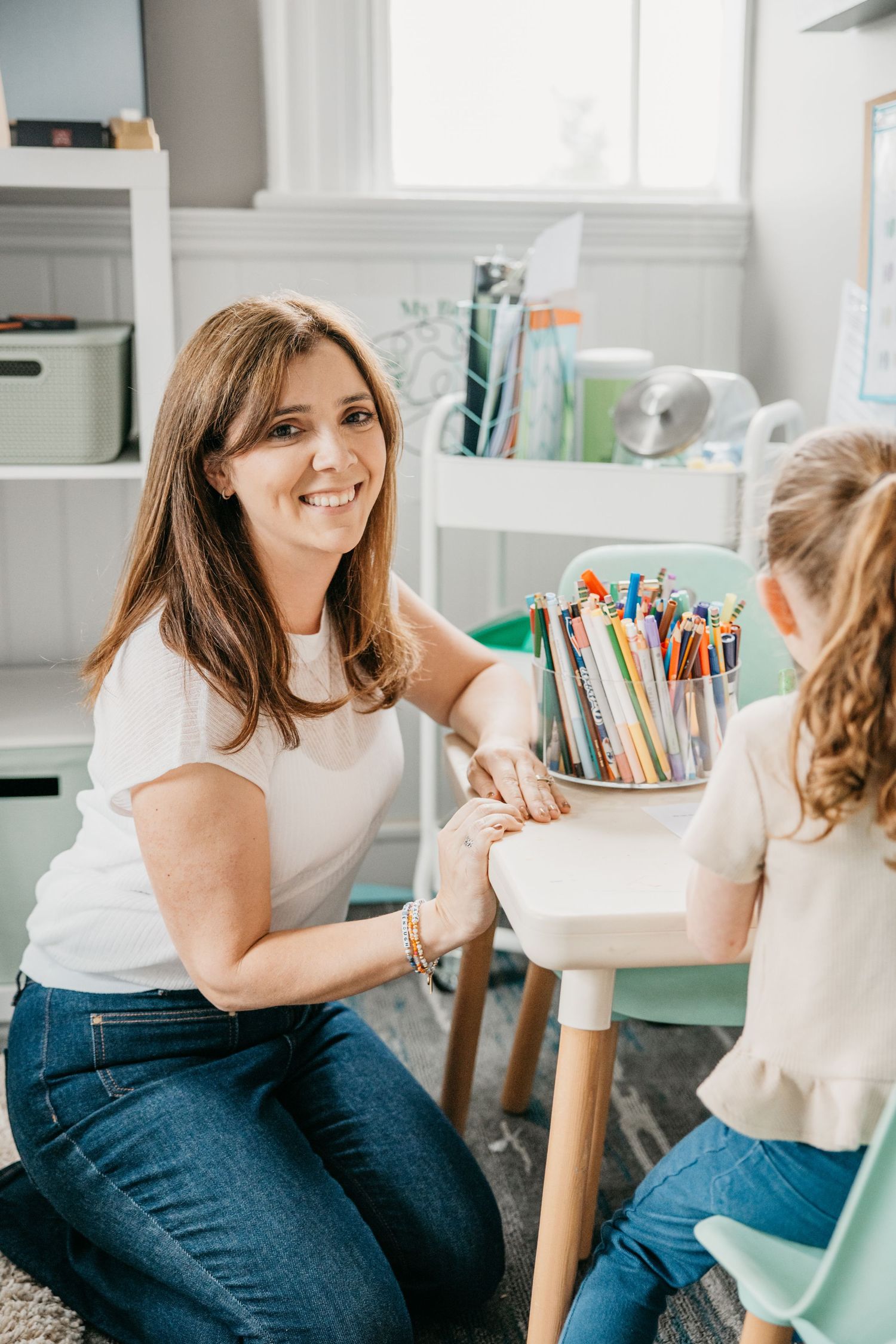 A smiling woman in a white top sits with a young girl at a small desk with colorful pencils in a bright playroom.