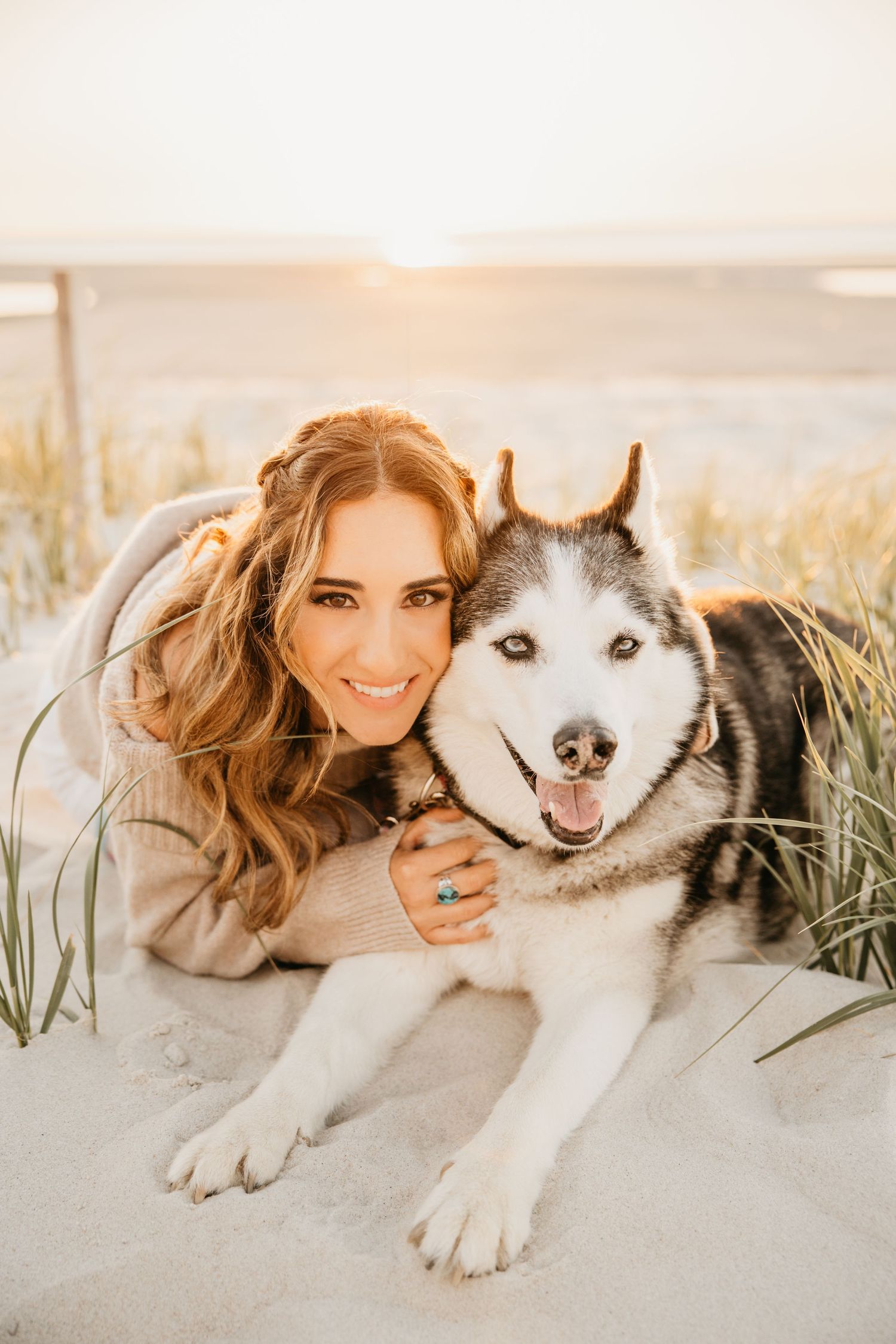 Woman smiling with a Siberian Husky dog on sandy beach at sunset, lying together in the sand dunes.