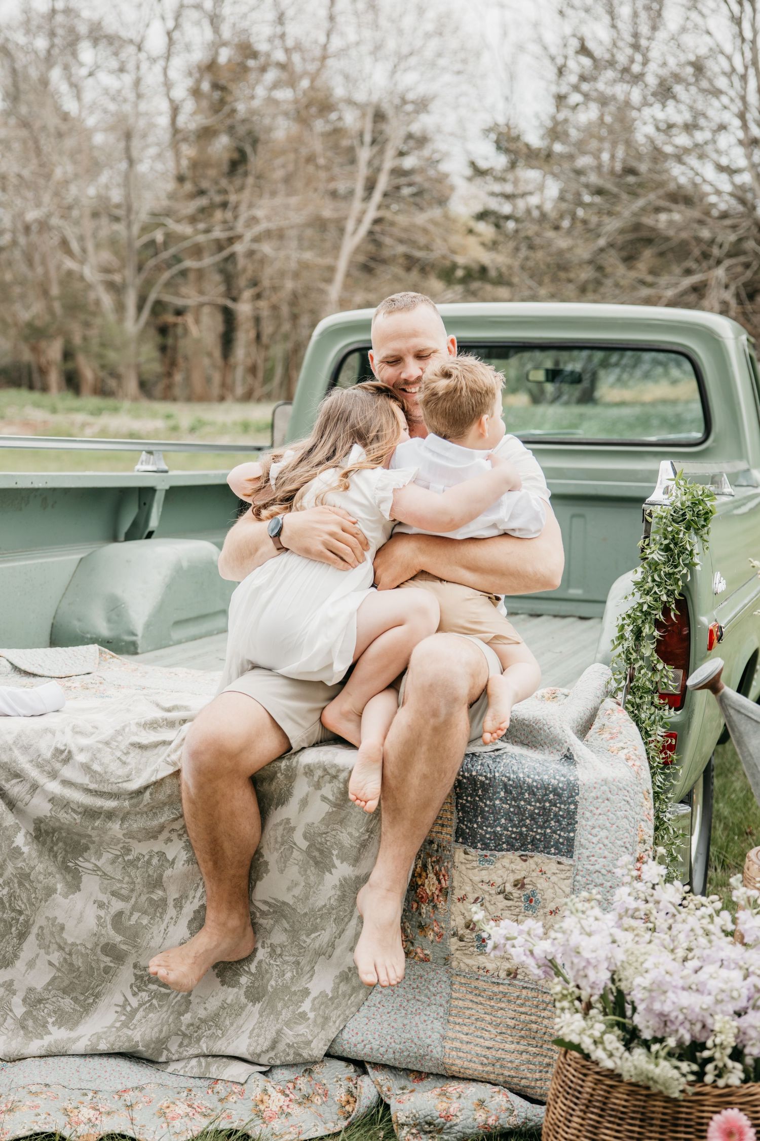 A smiling man sits on a vintage green truck bed, hugging two young children outdoors with floral decor nearby.
