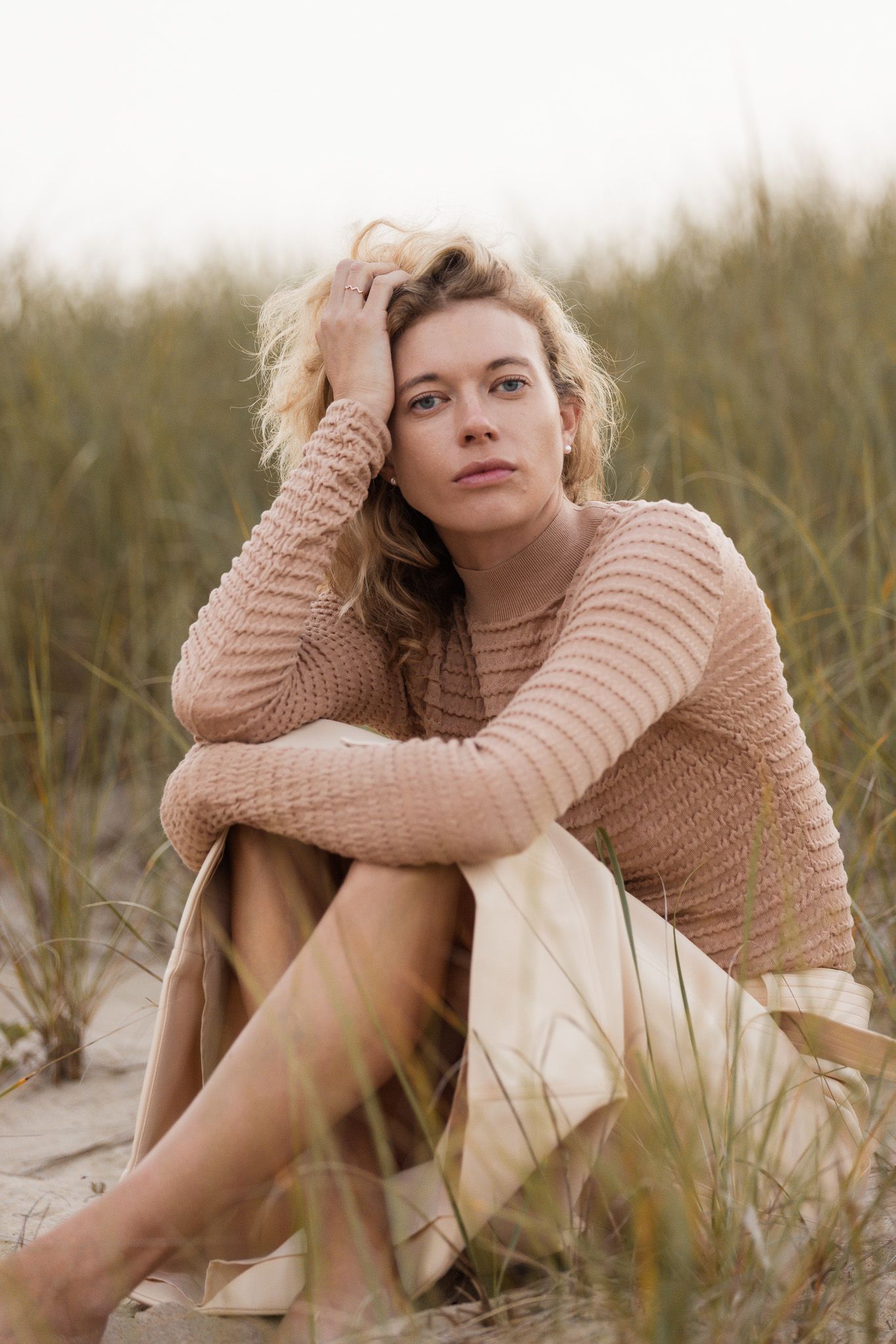 Person in beige sweater sitting thoughtfully among beach grass at sunset.