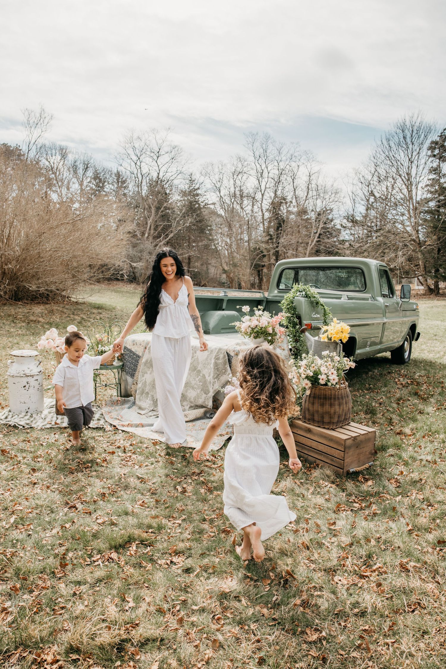 A woman in white dress plays with two young children near a vintage green truck decorated with flowers outdoors.