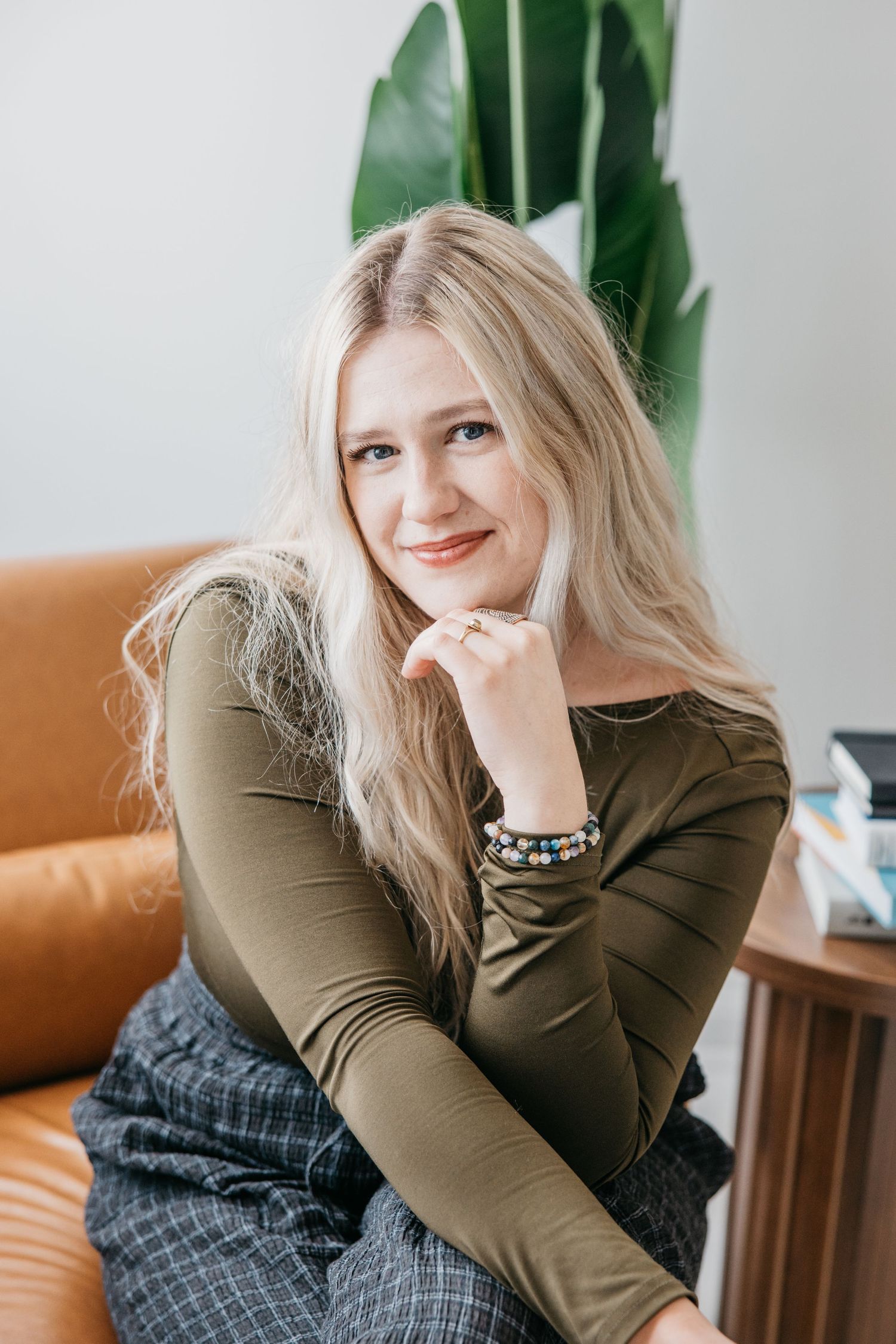 Smiling blonde woman in olive green top sitting on brown leather sofa with green plant in background.