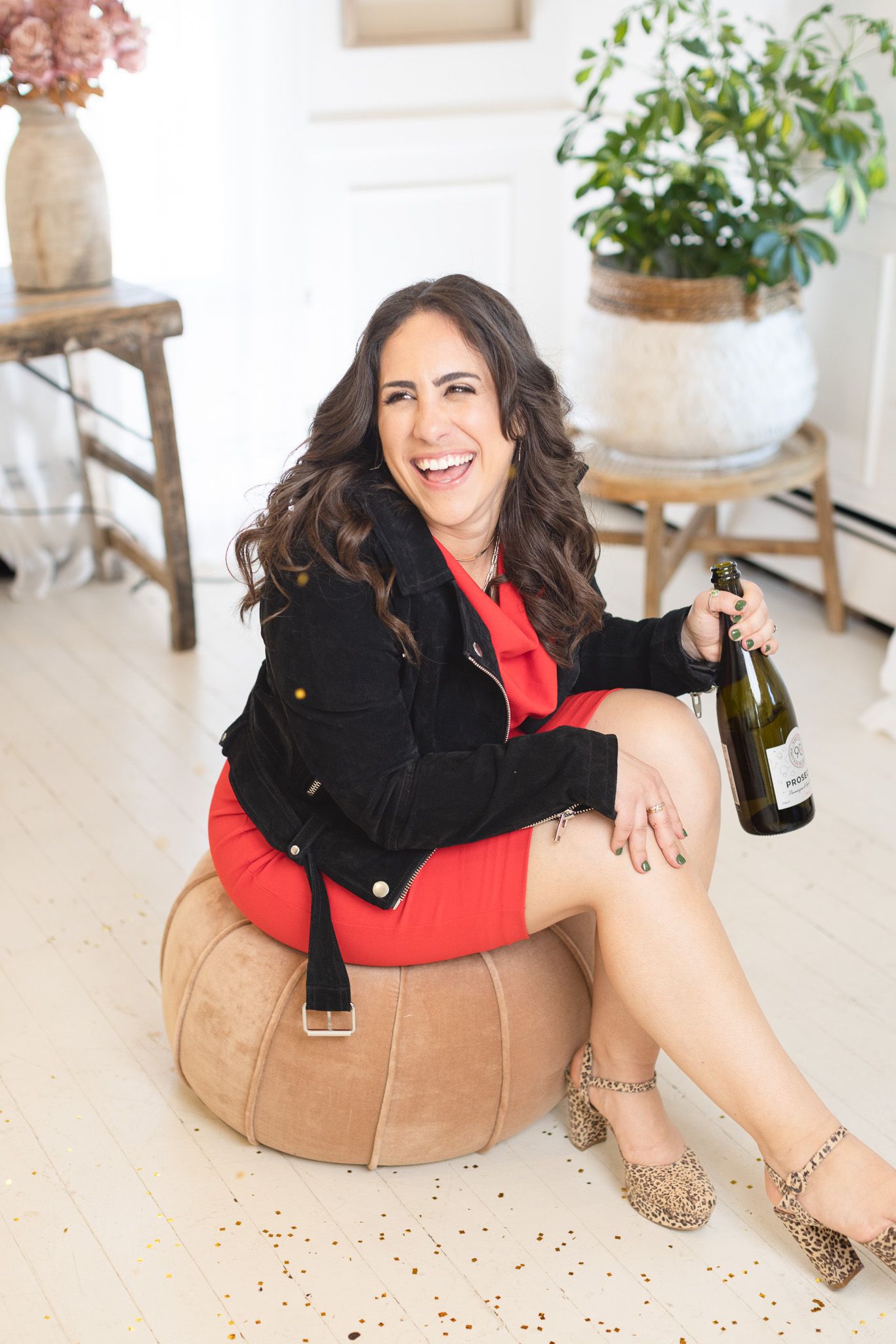 Casual lifestyle photos on tan pouf featuring black jacket and red top with champagne bottle.