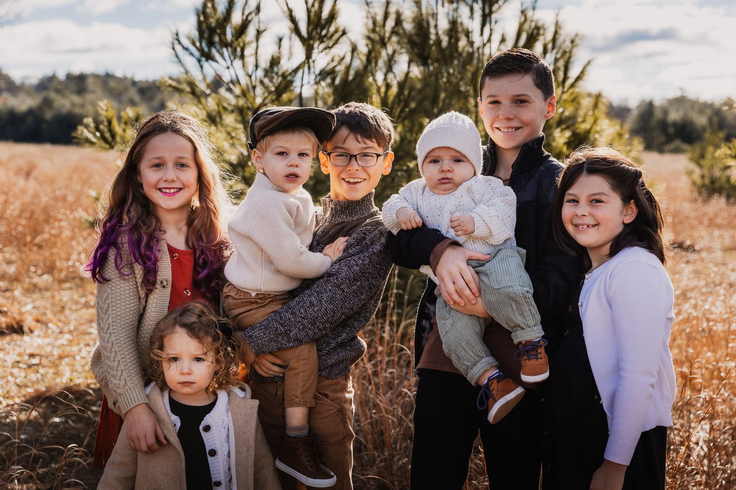 Family group photo in casual attire posed among pine trees in autumn setting.