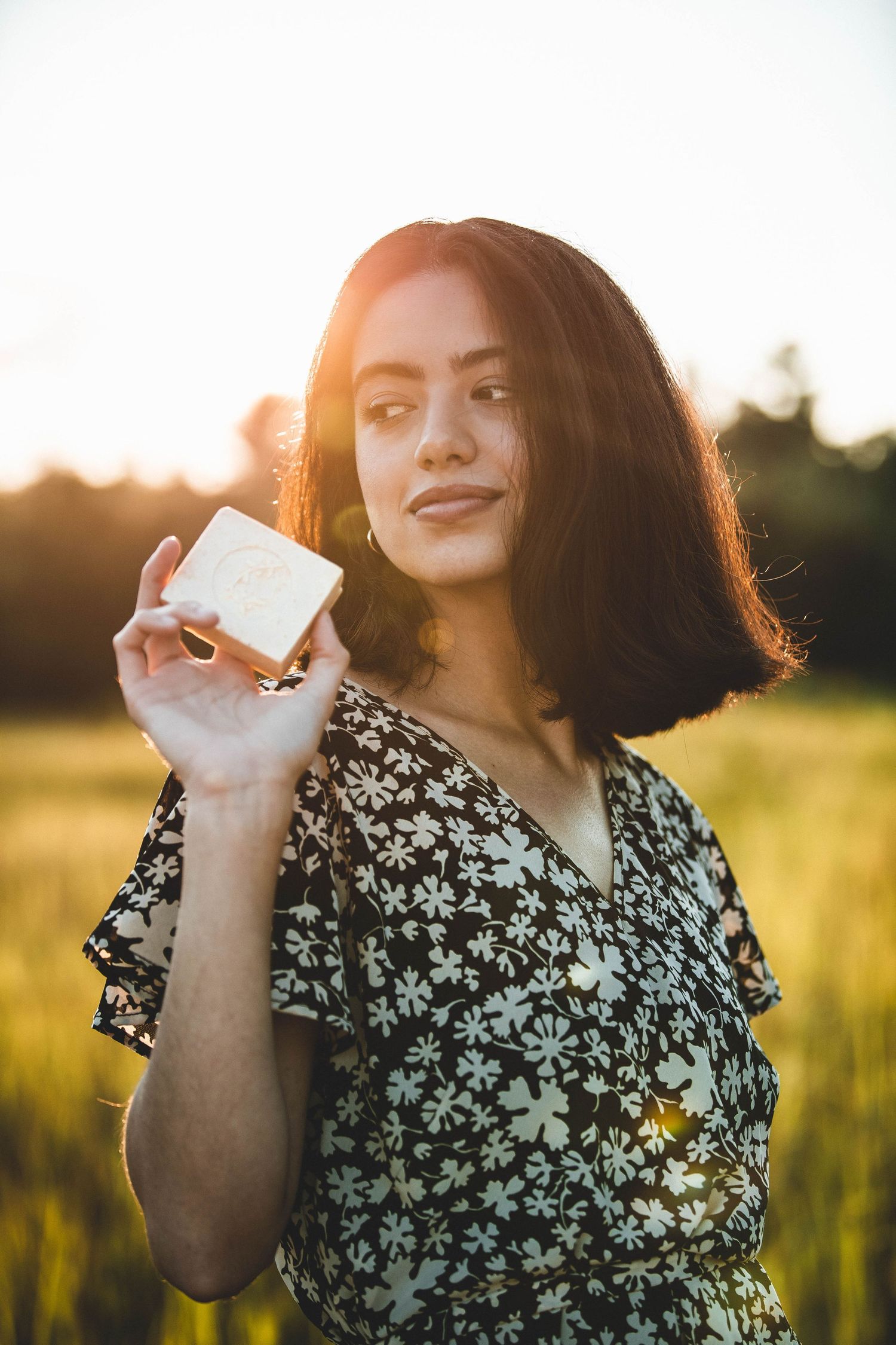 Person in floral dress holding product in sunlit field at sunset.