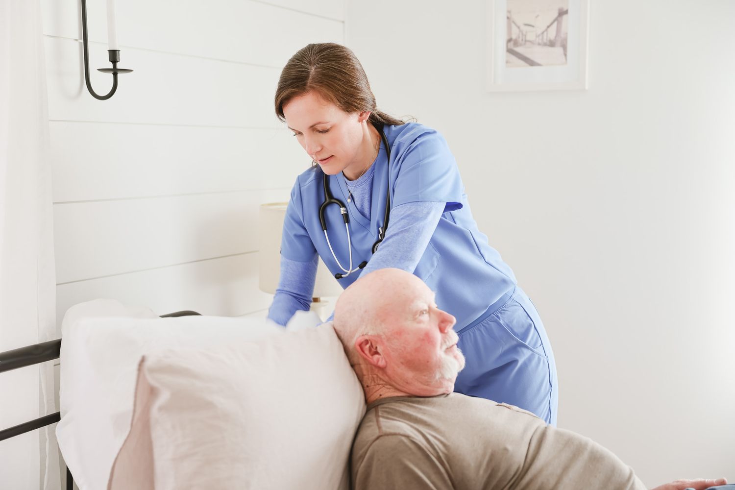 Female nurse in blue scrubs adjusting pillow for elderly male patient lying in bed at home.