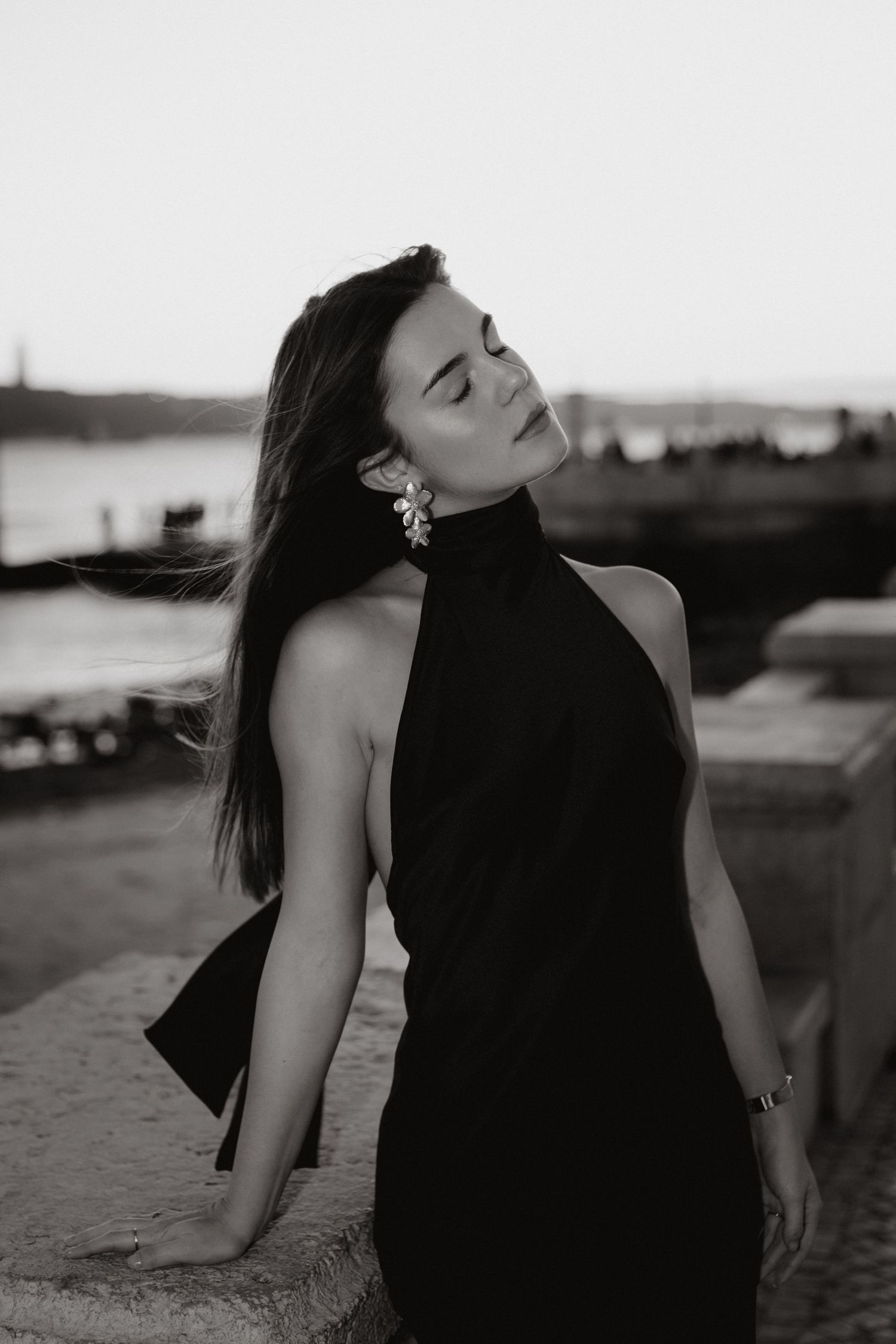Black and white portrait of a woman in a black halter dress with statement earrings posing near a river at dusk.