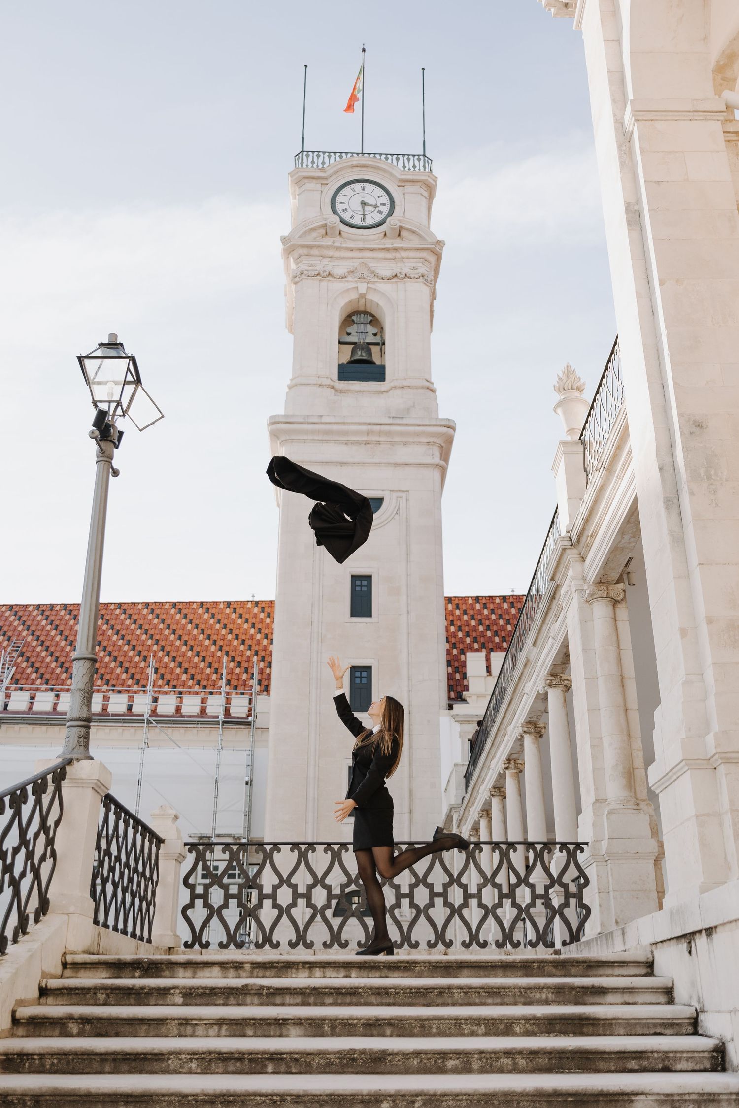 Woman tossing graduation cap in air on steps near historic clock tower building.
