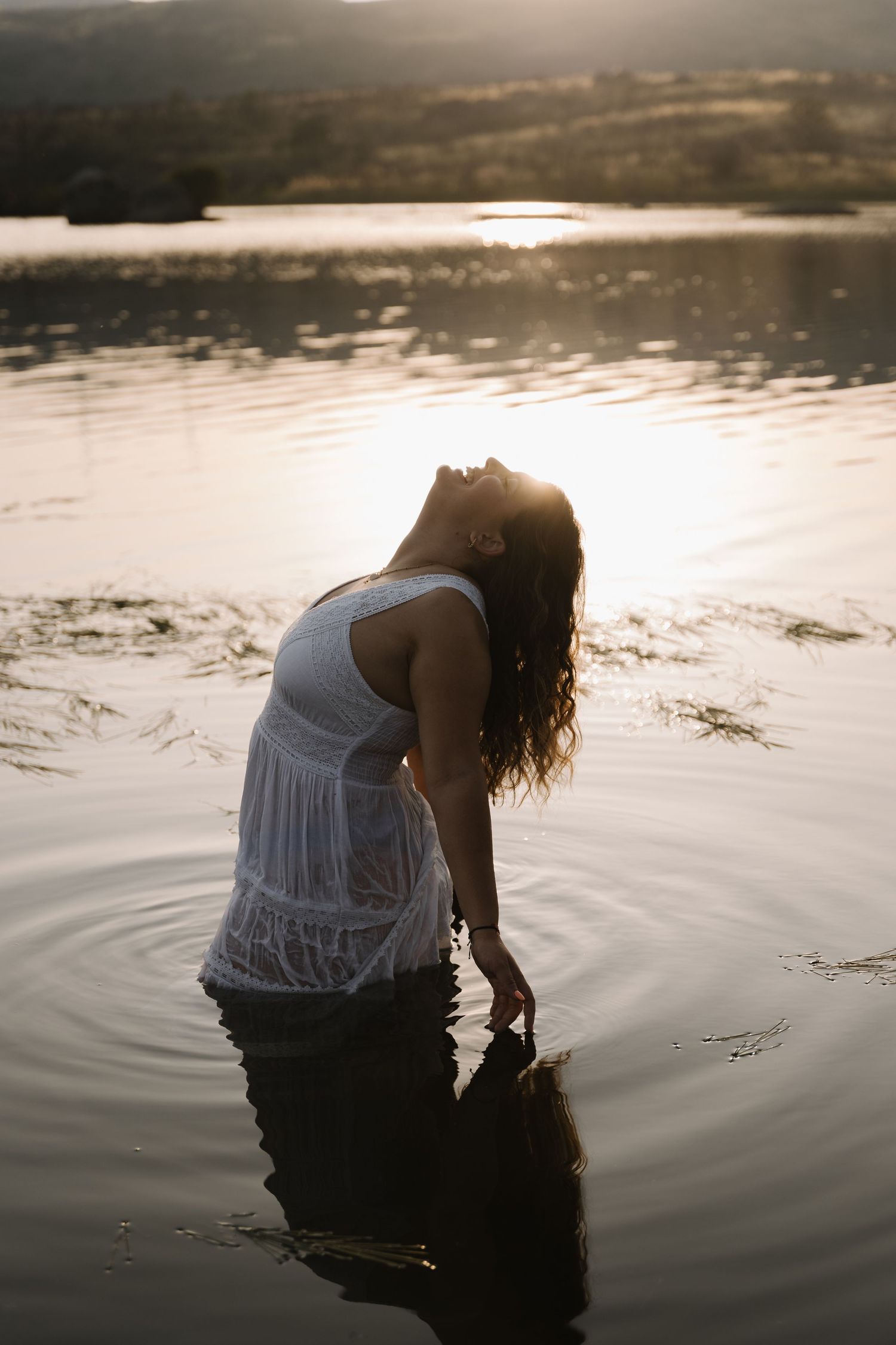 Woman in white dress standing in lake at golden sunset with rippling water reflections.