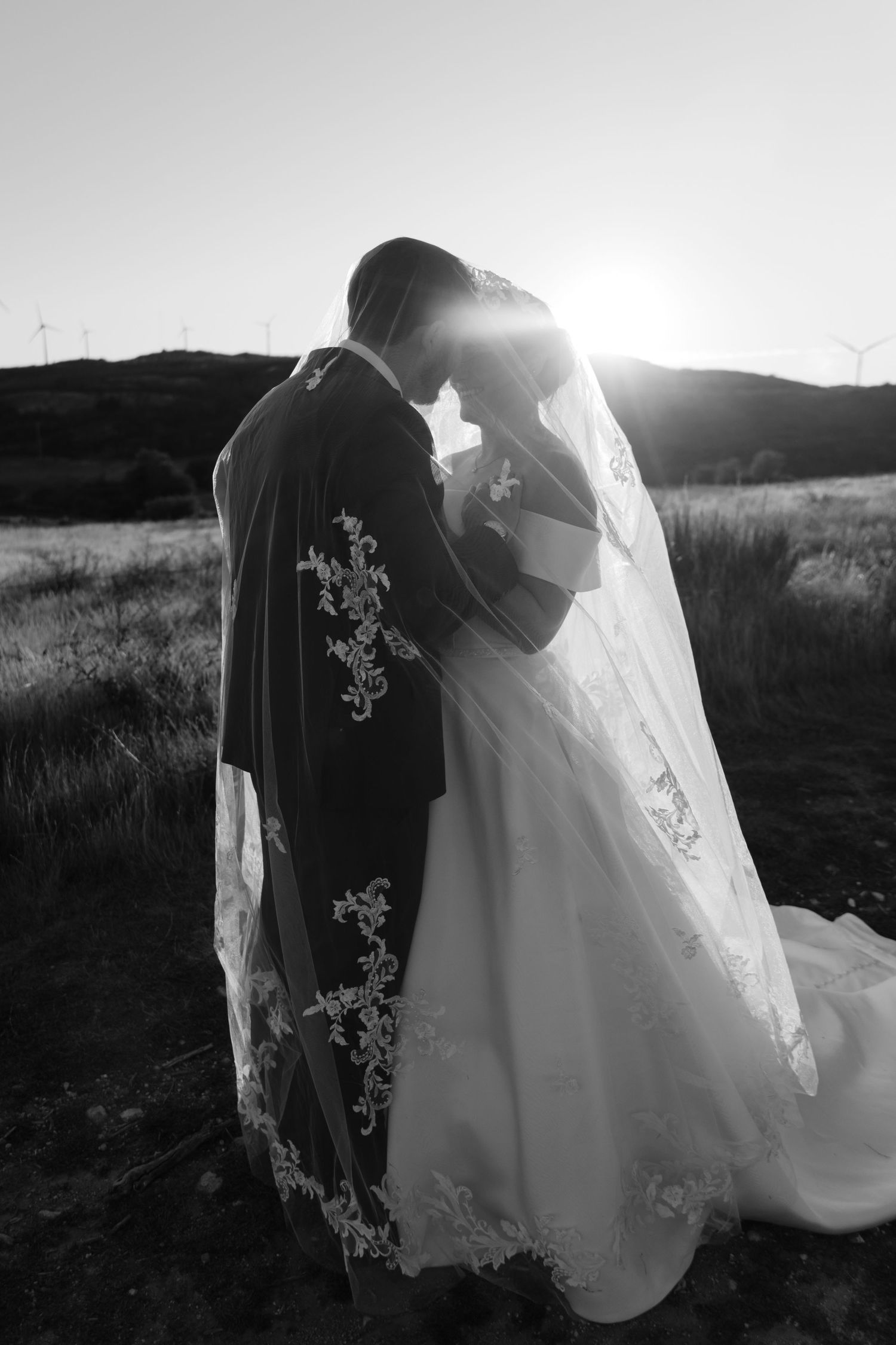Black and white photo of bride and groom embracing at sunset in an open field with wind turbines in the background.