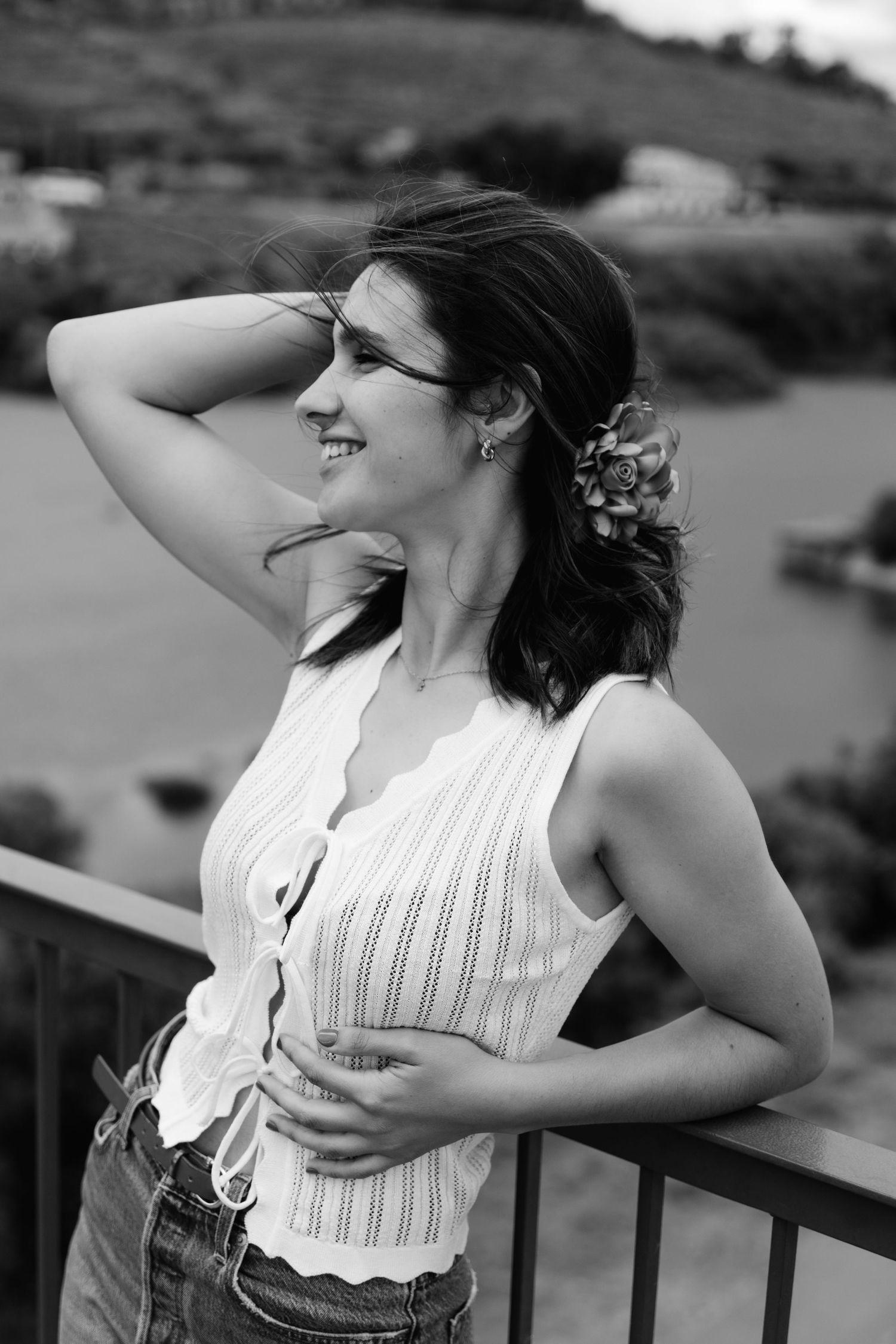 Black and white portrait of smiling brunette woman with flower in hair leaning on railing.