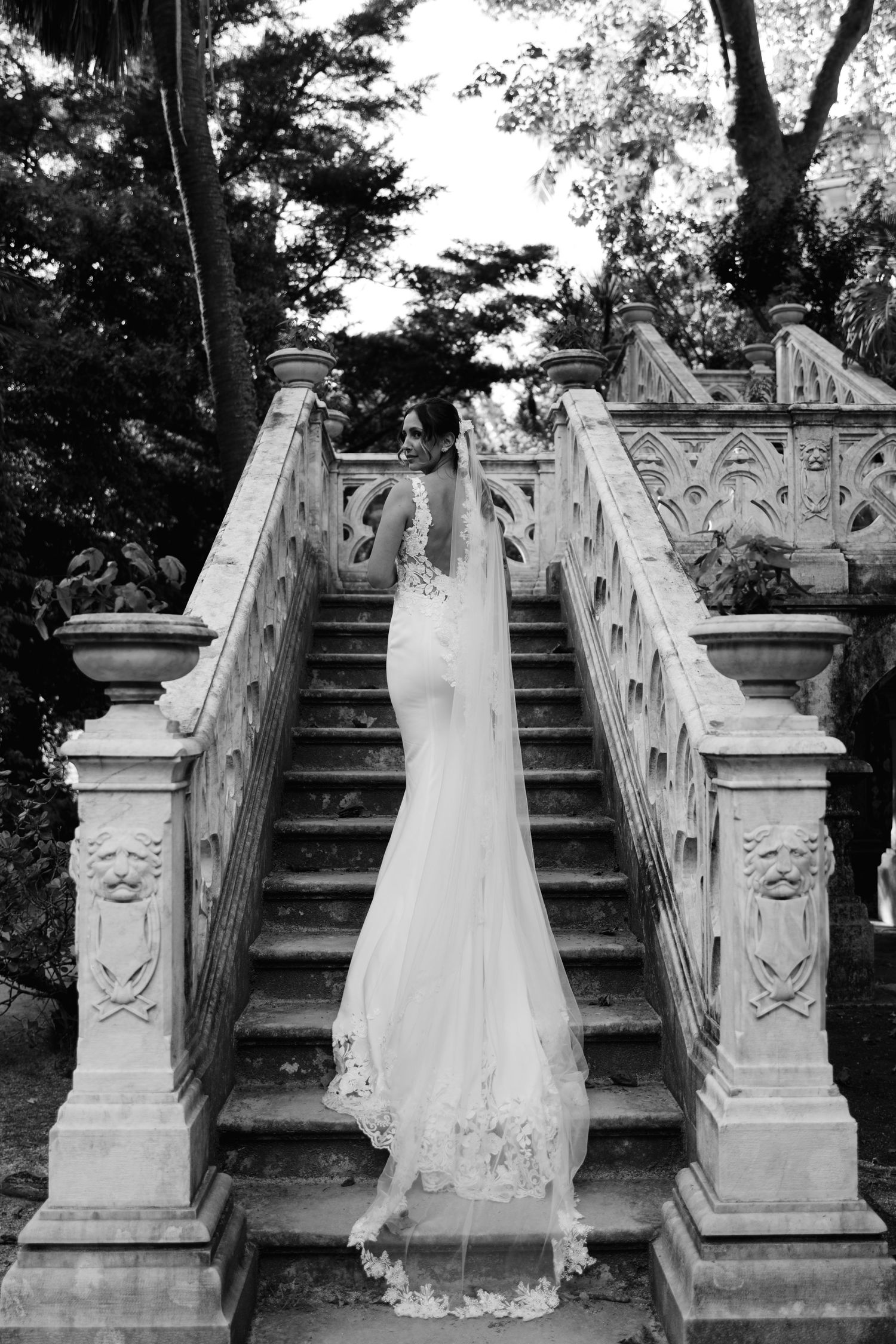 Bride in lace wedding gown ascending ornate stone staircase in black and white garden setting.