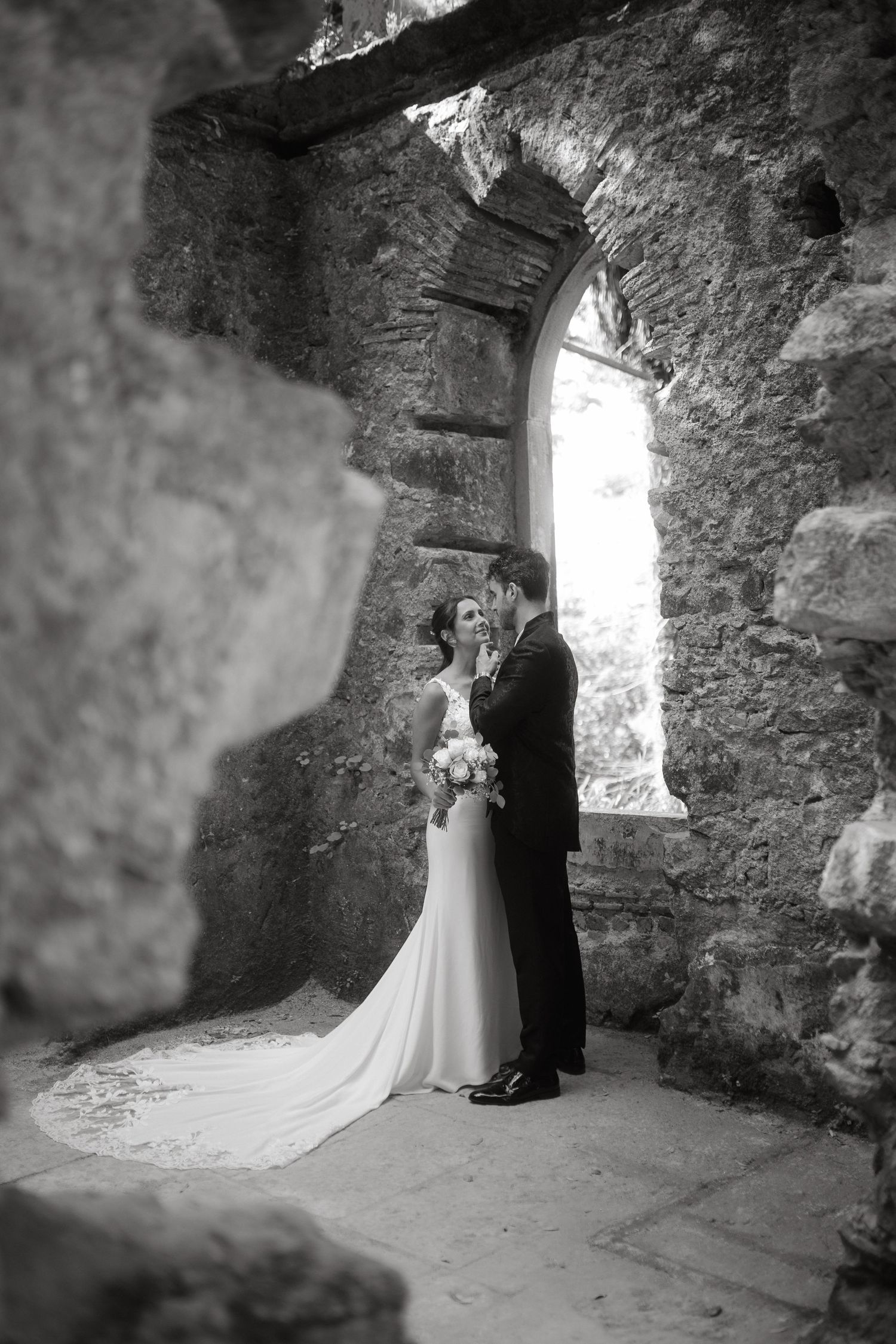 Bride and groom embracing beneath a gothic stone arch in black and white wedding photography.