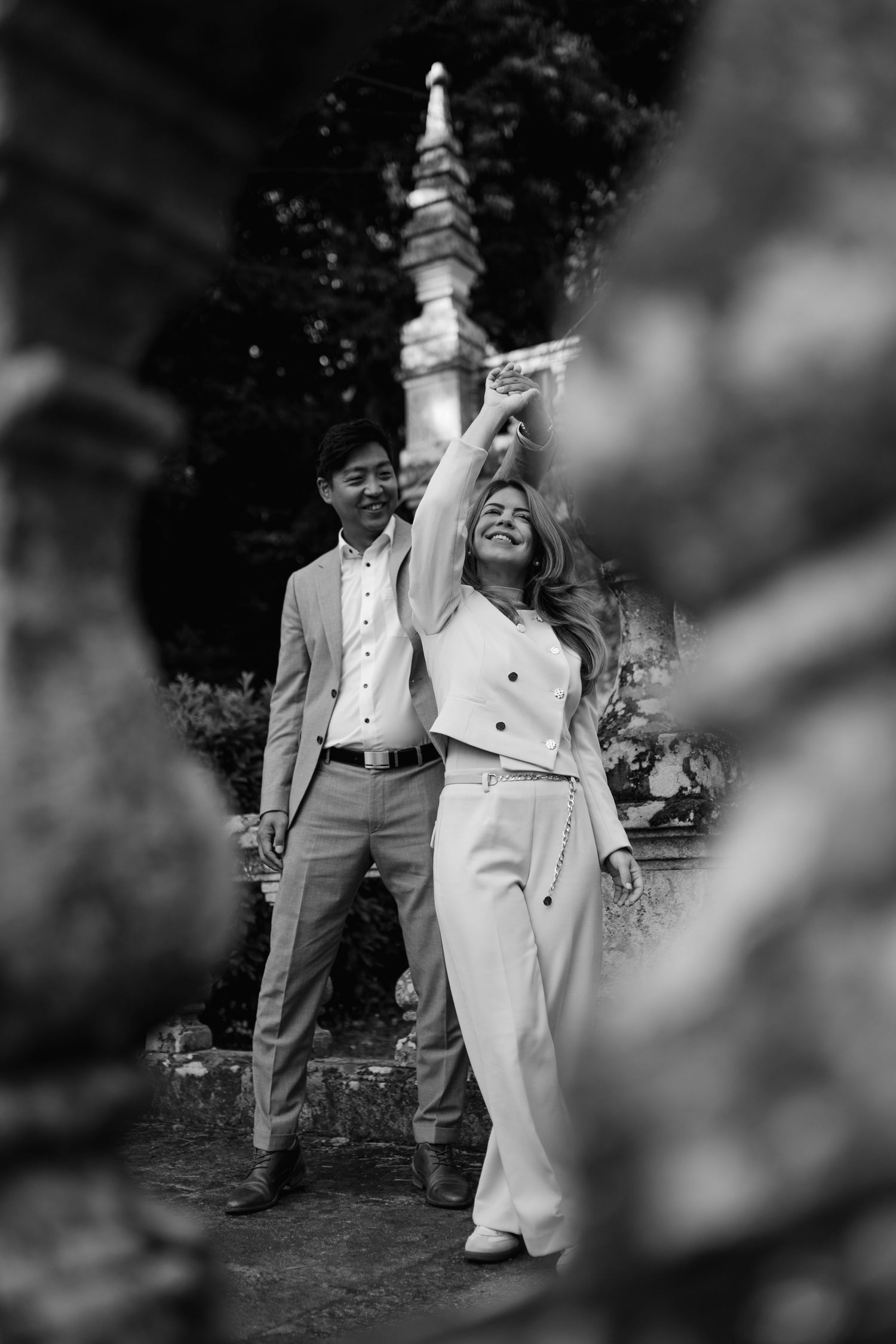 Black and white photo of a couple posing near ornate stone architecture, woman in white outfit raising her hand. Pre-wedding photoshoot