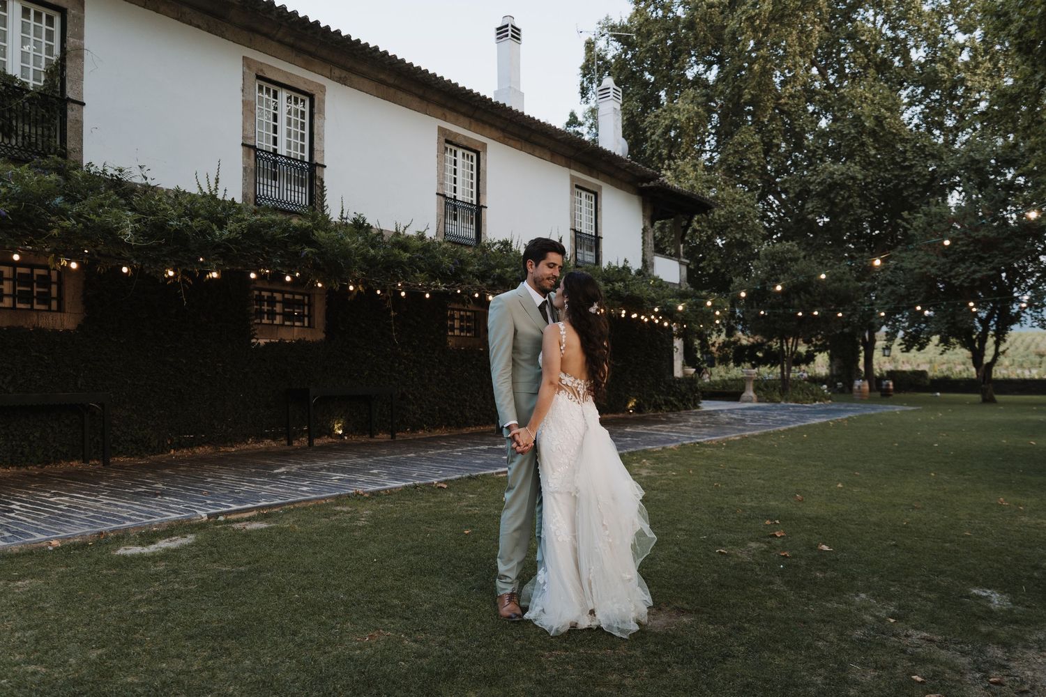 Wedding couple embracing on green lawn in front of elegant venue with string lights at dusk.