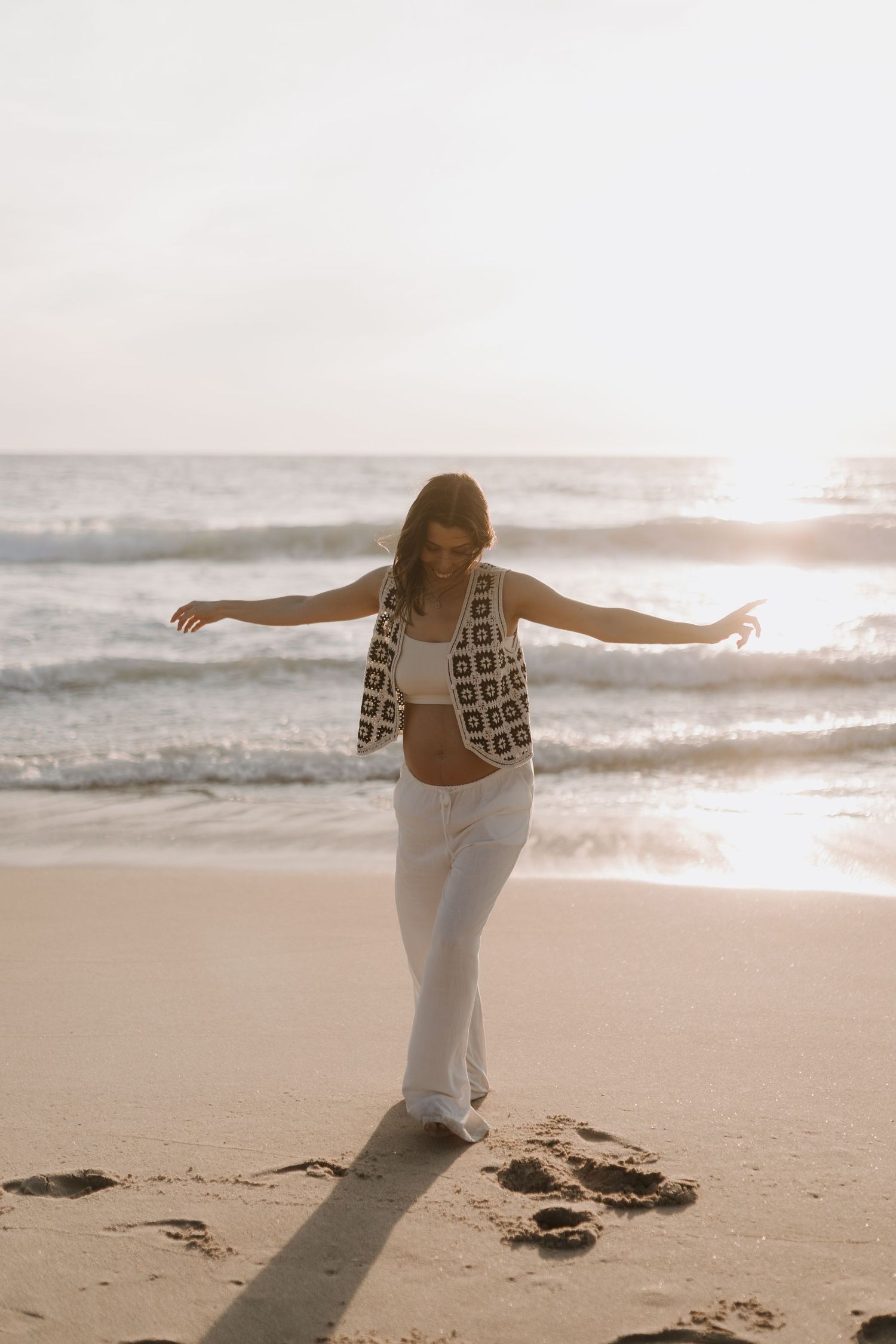 Pregnant woman in crochet top and white pants balancing on sunny beach with arms outstretched.
