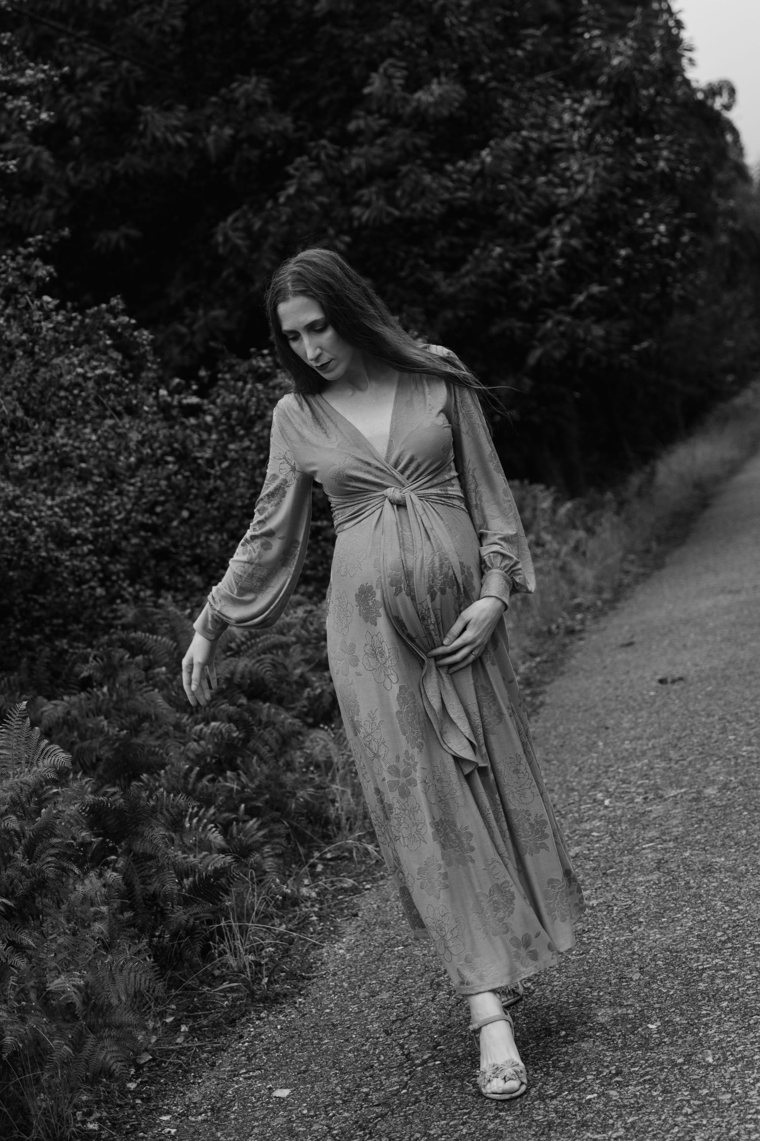 Elegant pregnant woman in flowy dress walking on outdoor path surrounded by lush greenery.