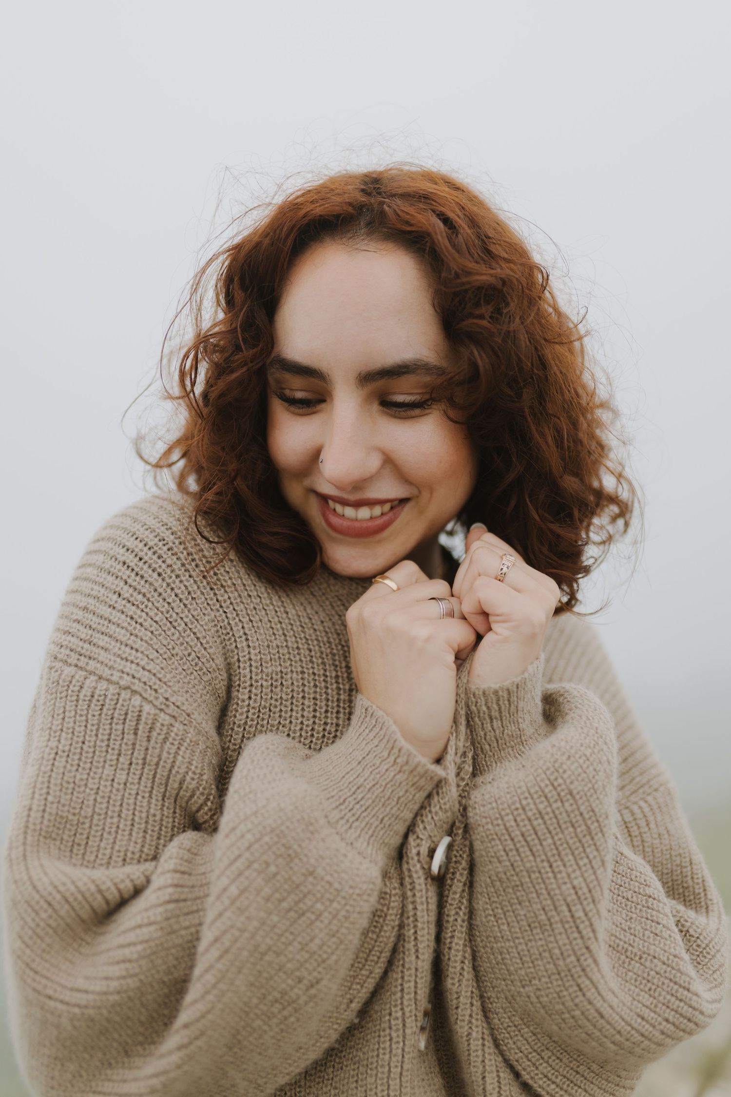 Smiling woman with curly red hair wearing a cozy beige knit cardigan, holding collar with ringed fingers. Fog day