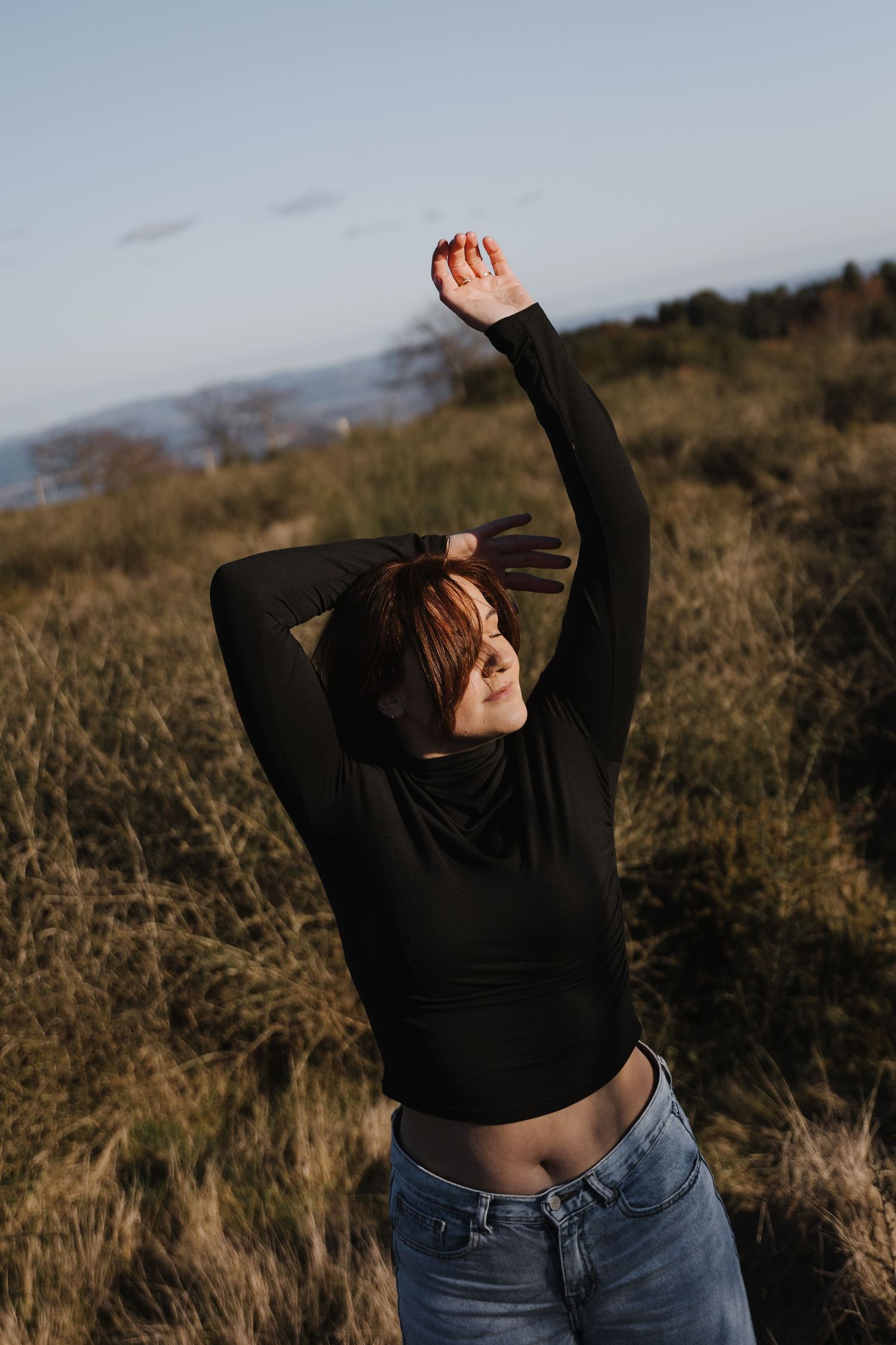 Young woman in black crop top and jeans stretching arms overhead in a golden sunlit grassy field.