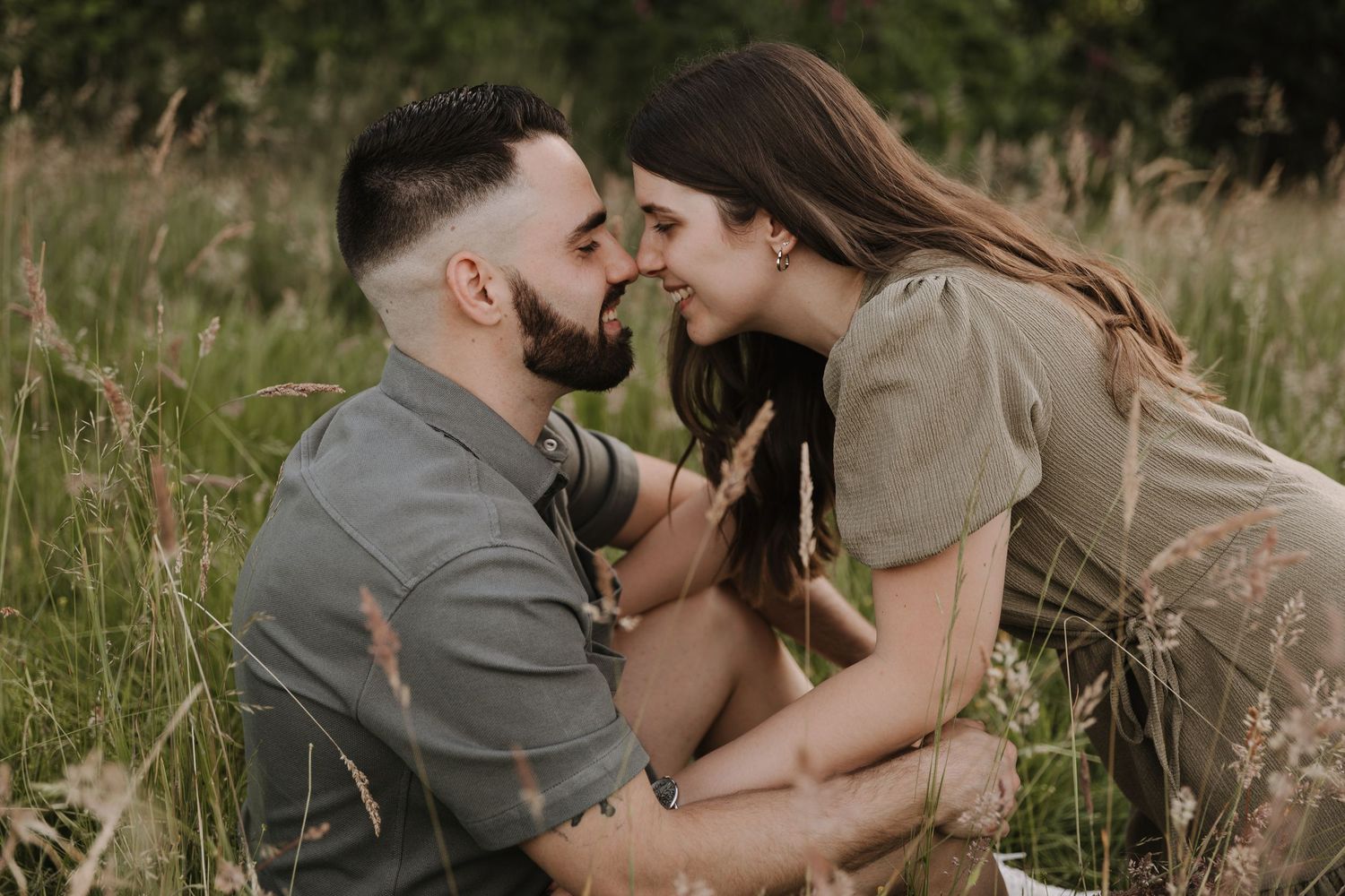 Romantic couple sitting in tall grass field, smiling and touching noses affectionately.