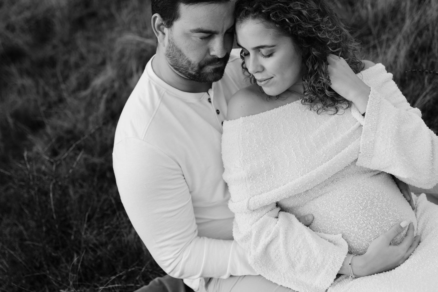 Black and white maternity photo of couple embracing outdoors, man tenderly holding pregnant woman.