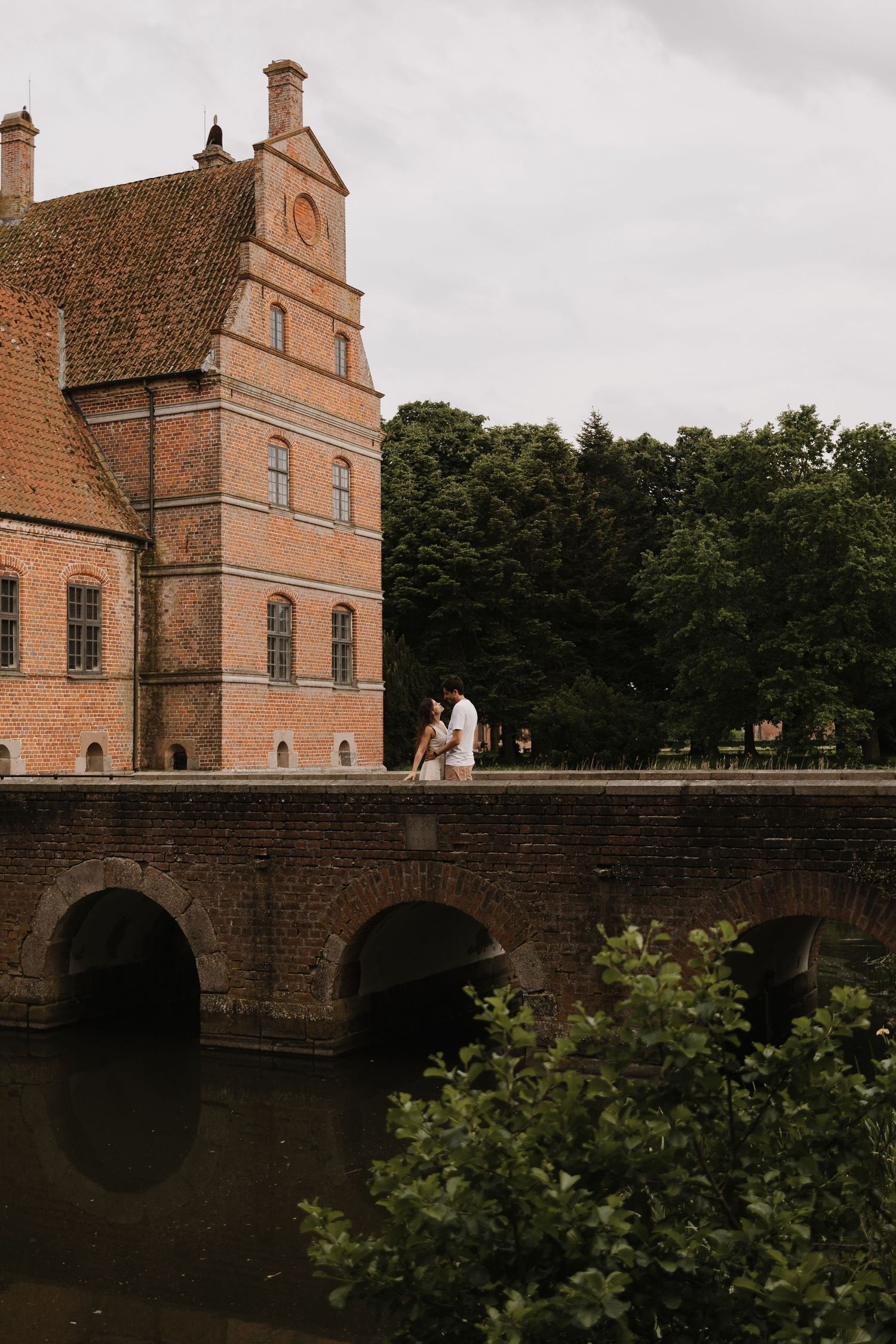Couple sharing a romantic moment on a stone bridge beside a historic brick castle near a serene moat.