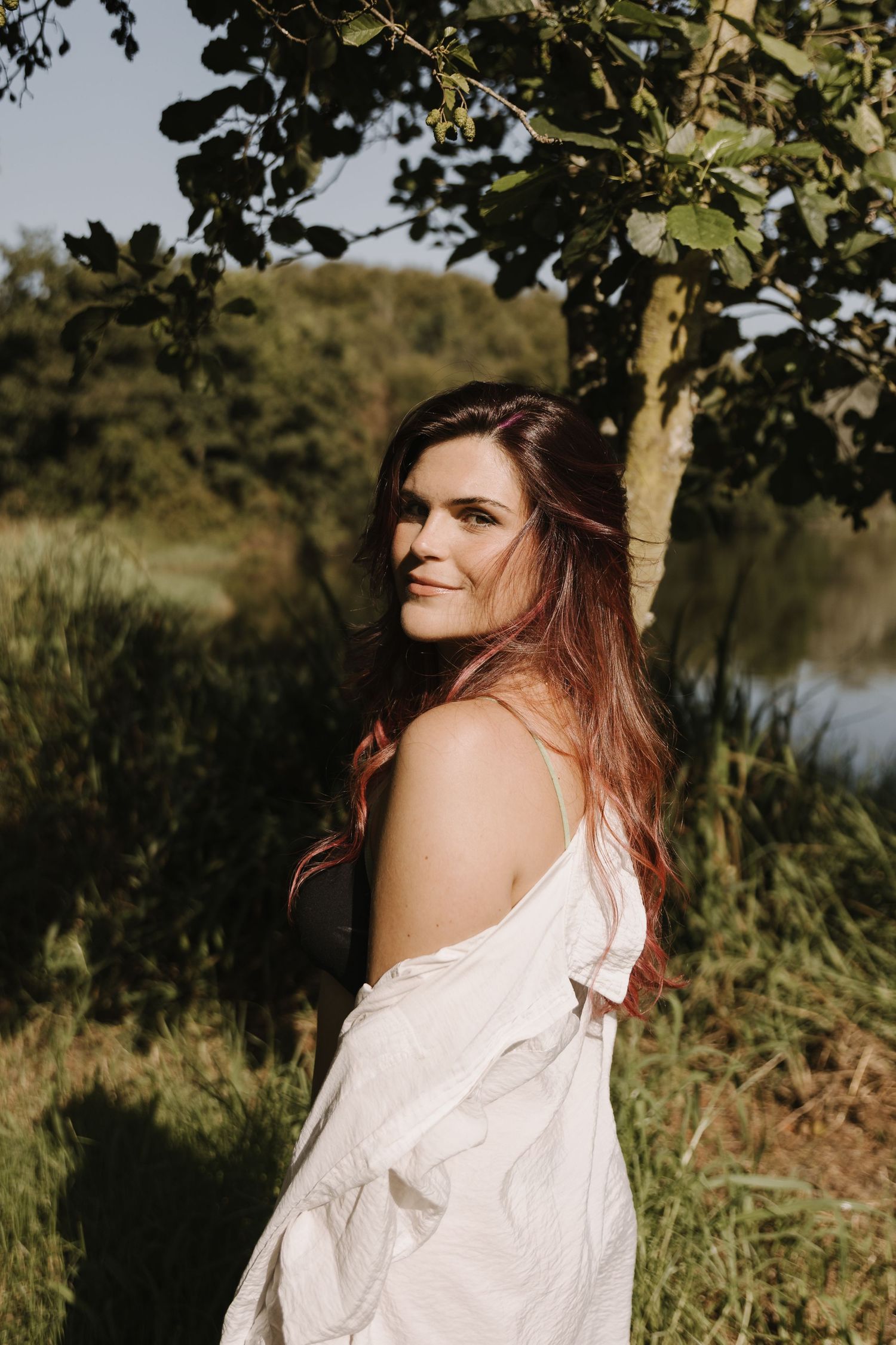 A woman with red hair in a white shirt smiles softly while standing near a tree by a peaceful lake.