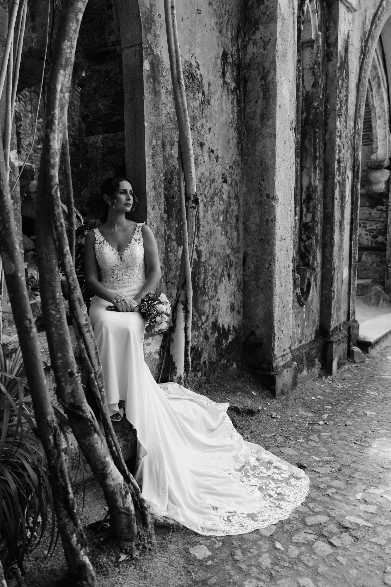 Bride in elegant lace wedding gown seated by ancient stone wall with tree roots in black and white.