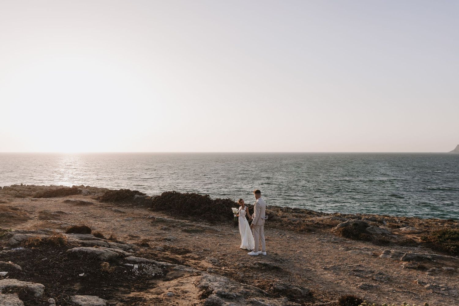Bride and groom holding hands on rocky coastal cliff with ocean horizon at sunset. Wedding photoshoot. Destination Wedding