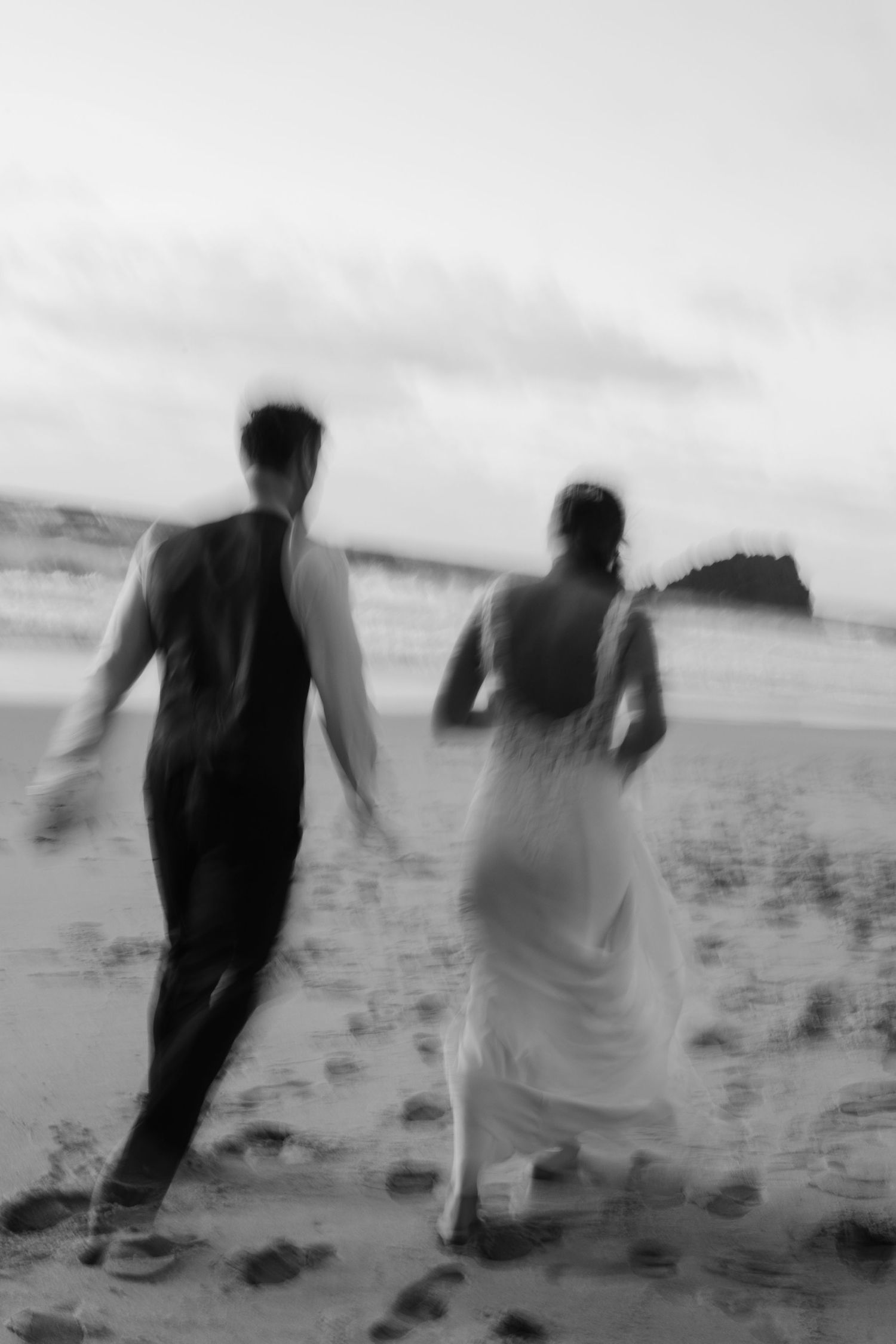 Blurred black and white photo of bride and groom running on beach, leaving footprints in sand. Trash the dress