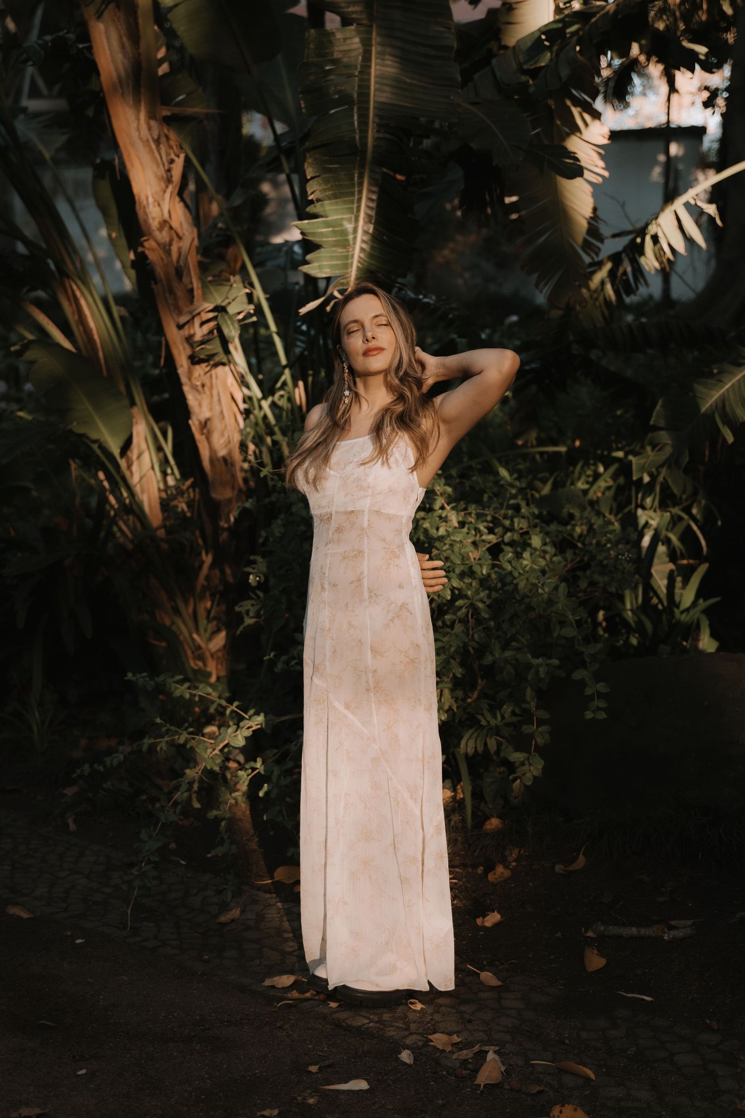 A woman in a flowing white maxi dress stands gracefully among lush tropical trees in golden evening light.