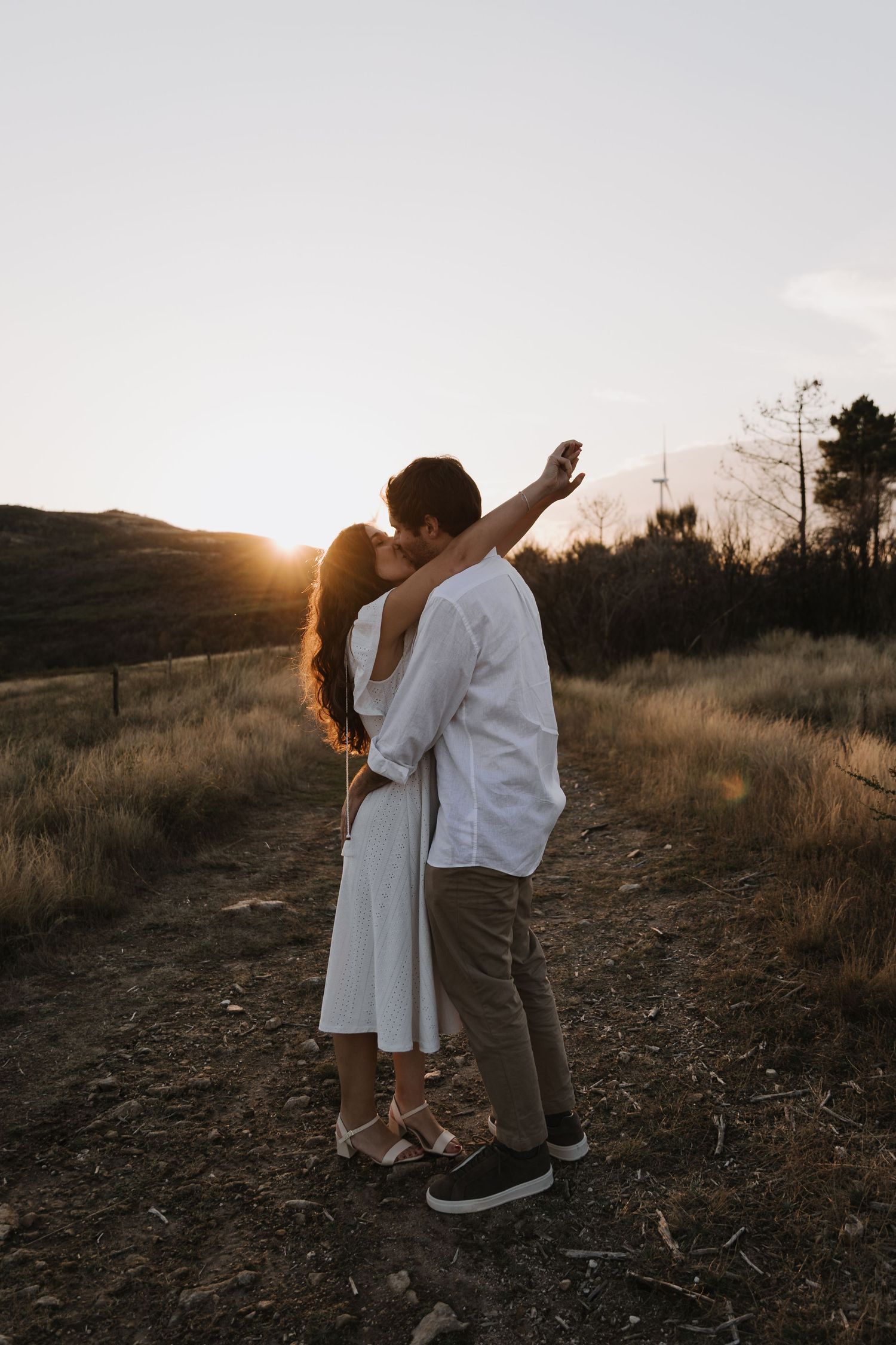 Couple embracing and kissing at golden hour sunset in an open field with warm backlit glow.