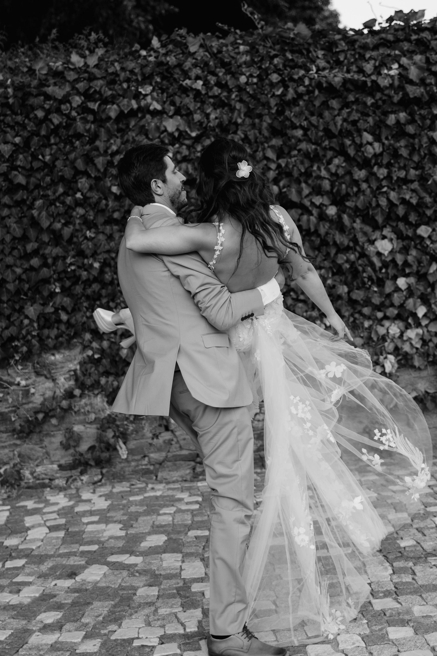 Black and white photo of a groom lifting his bride in a wedding dress outdoors against an ivy-covered wall.