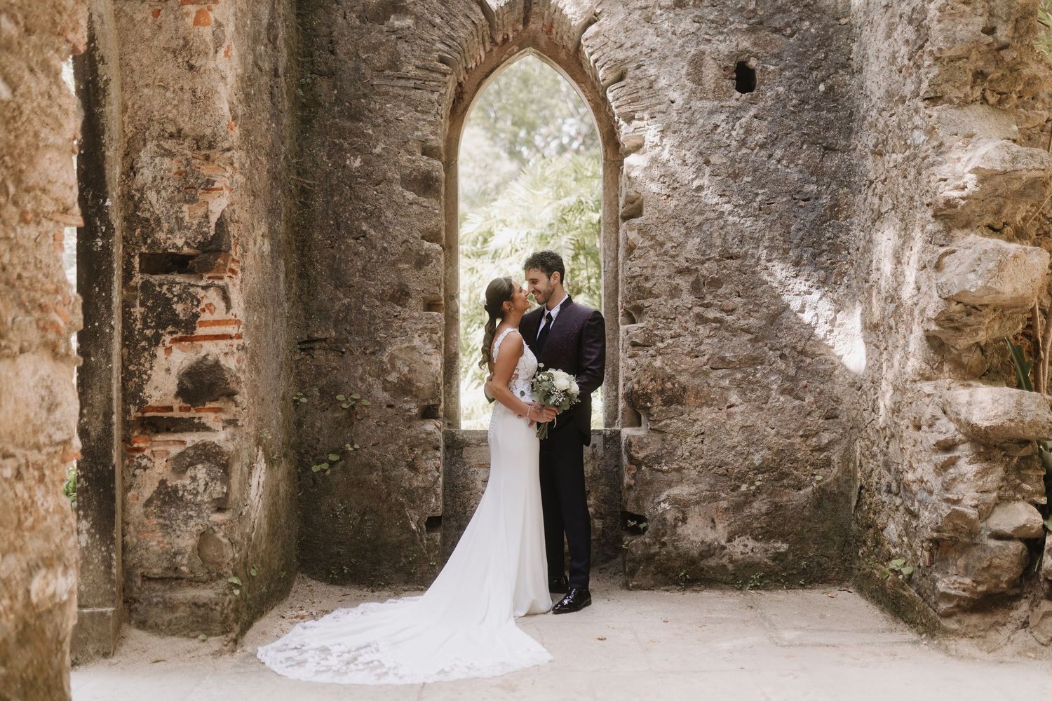Bride and groom sharing romantic kiss in stunning stone archway ruins at outdoor wedding venue.
