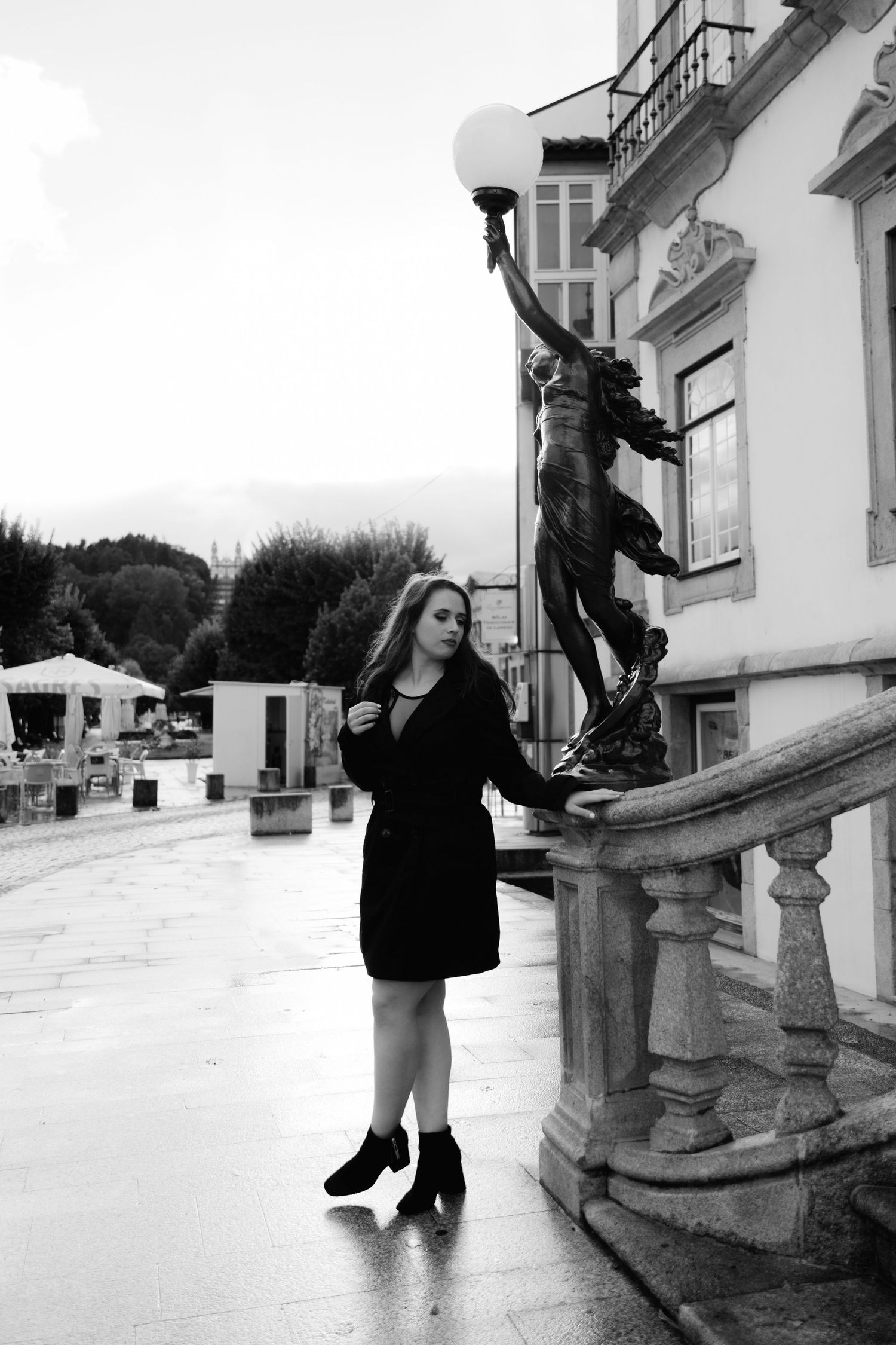 Black and white photo of a young woman in a black dress posing beside an ornate lamp statue outside a historic building.