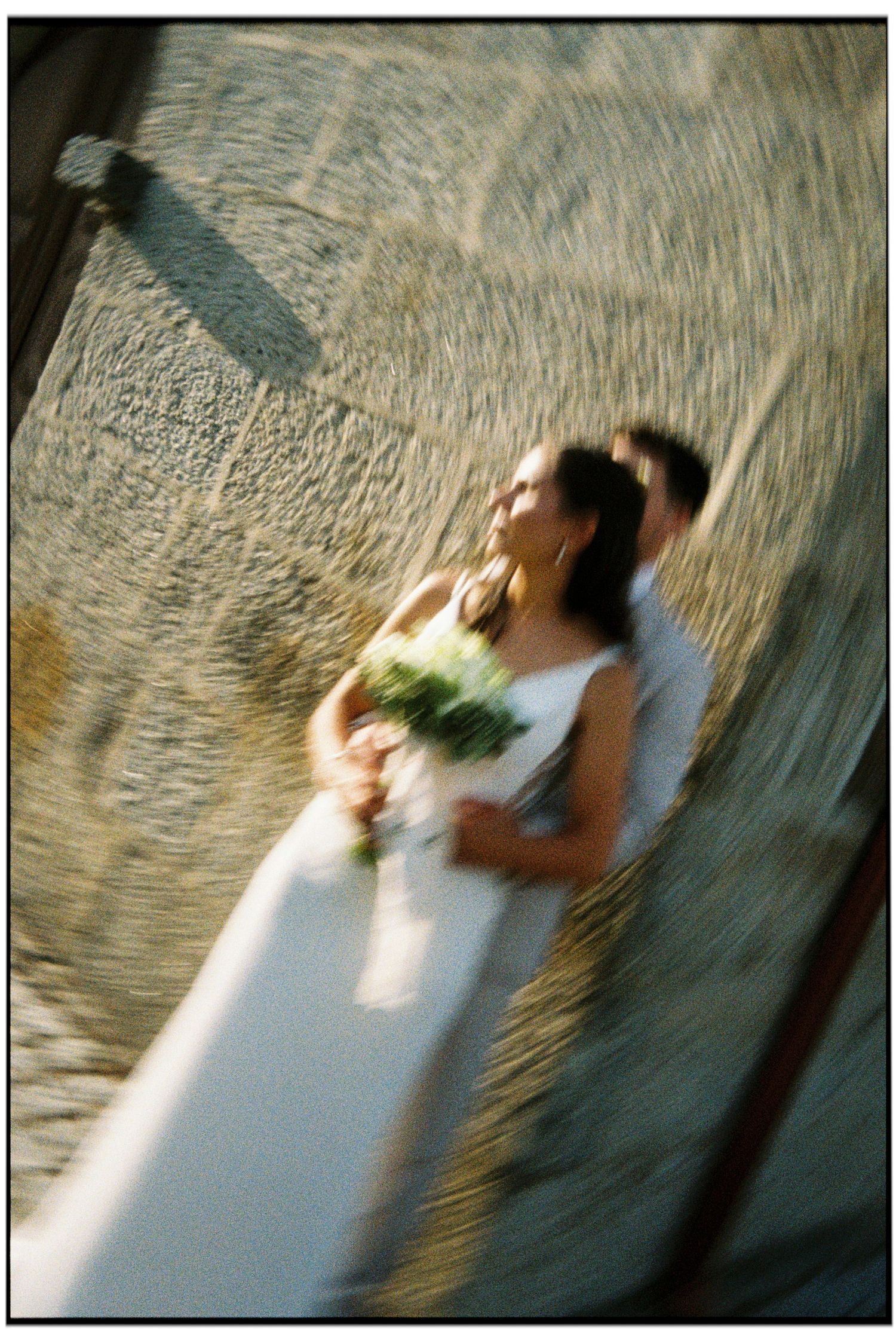 Bride in white gown holding bouquet stands with groom against stone wall in artistic tilted wedding photo. Film Photography. Film photograph