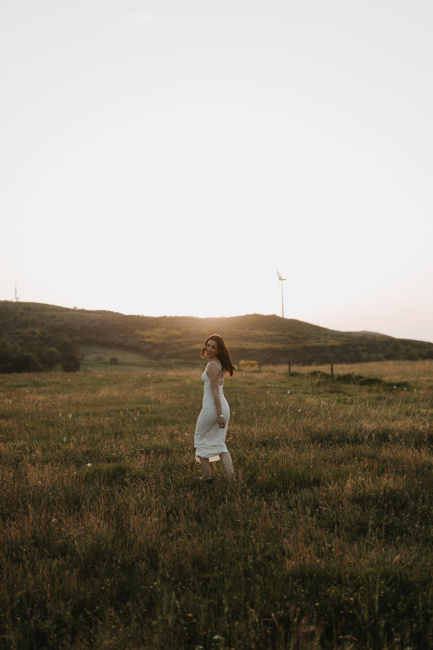 A woman in a white dress walks through a sunlit grassy field with wind turbines and hills in the background.
