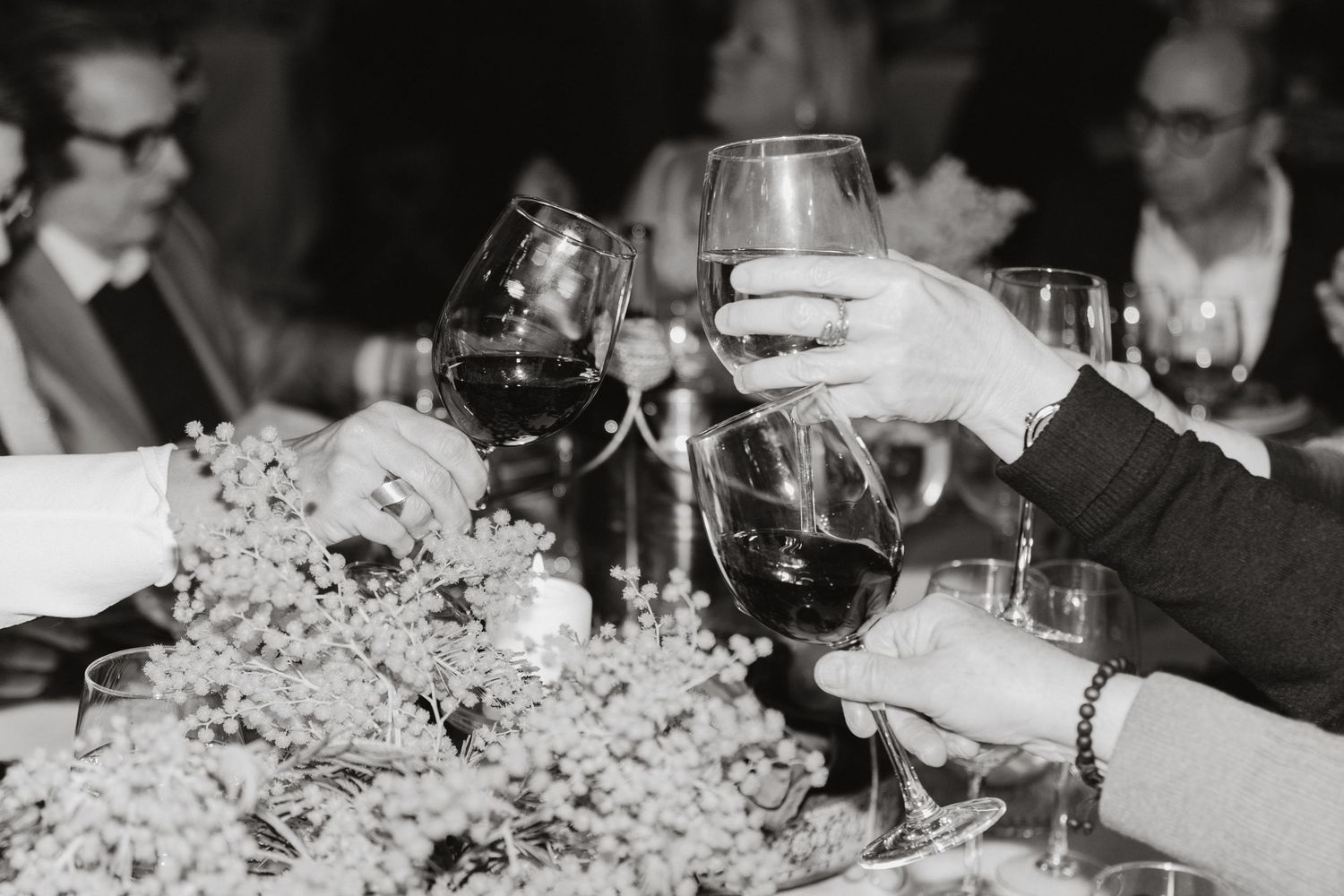 Black and white photo of friends toasting with wine glasses over floral centerpiece.