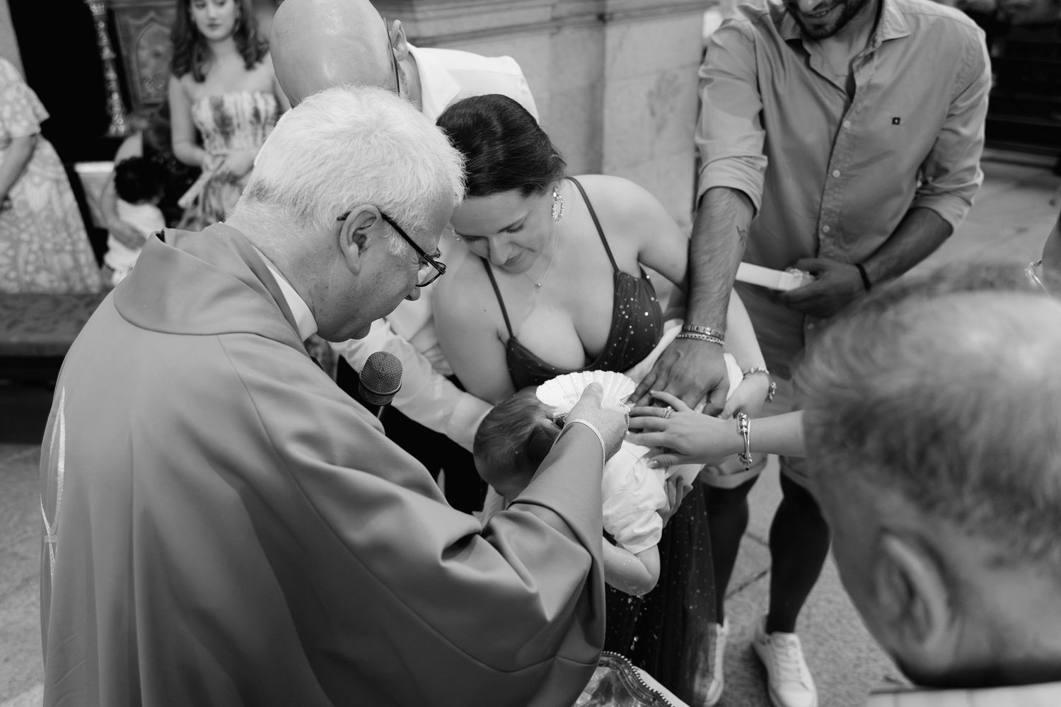 Black and white photo of a priest performing a baptism ceremony with parents and guests gathered closely.